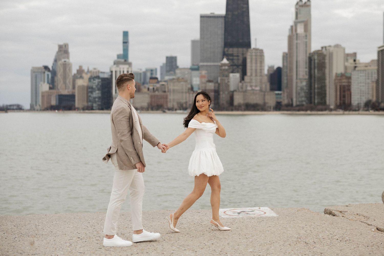 Couple holding hands on Chicago lakefront pier with city skyline backdrop, woman in white dress, man in beige suit.