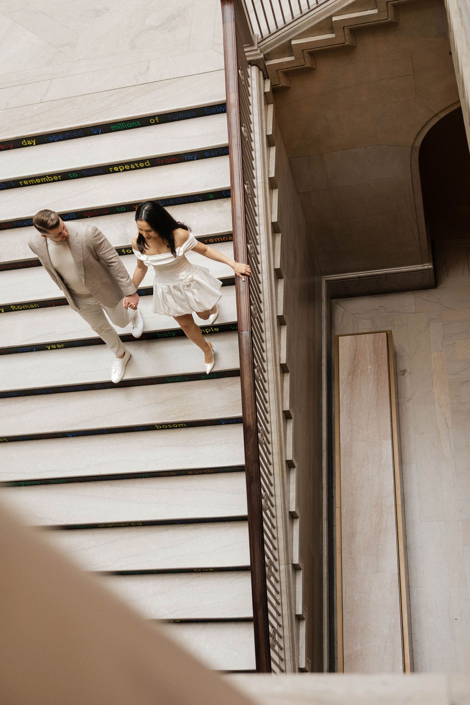 Aerial view of a couple holding hands walking up elegant stairs, woman in white dress and man in beige suit.