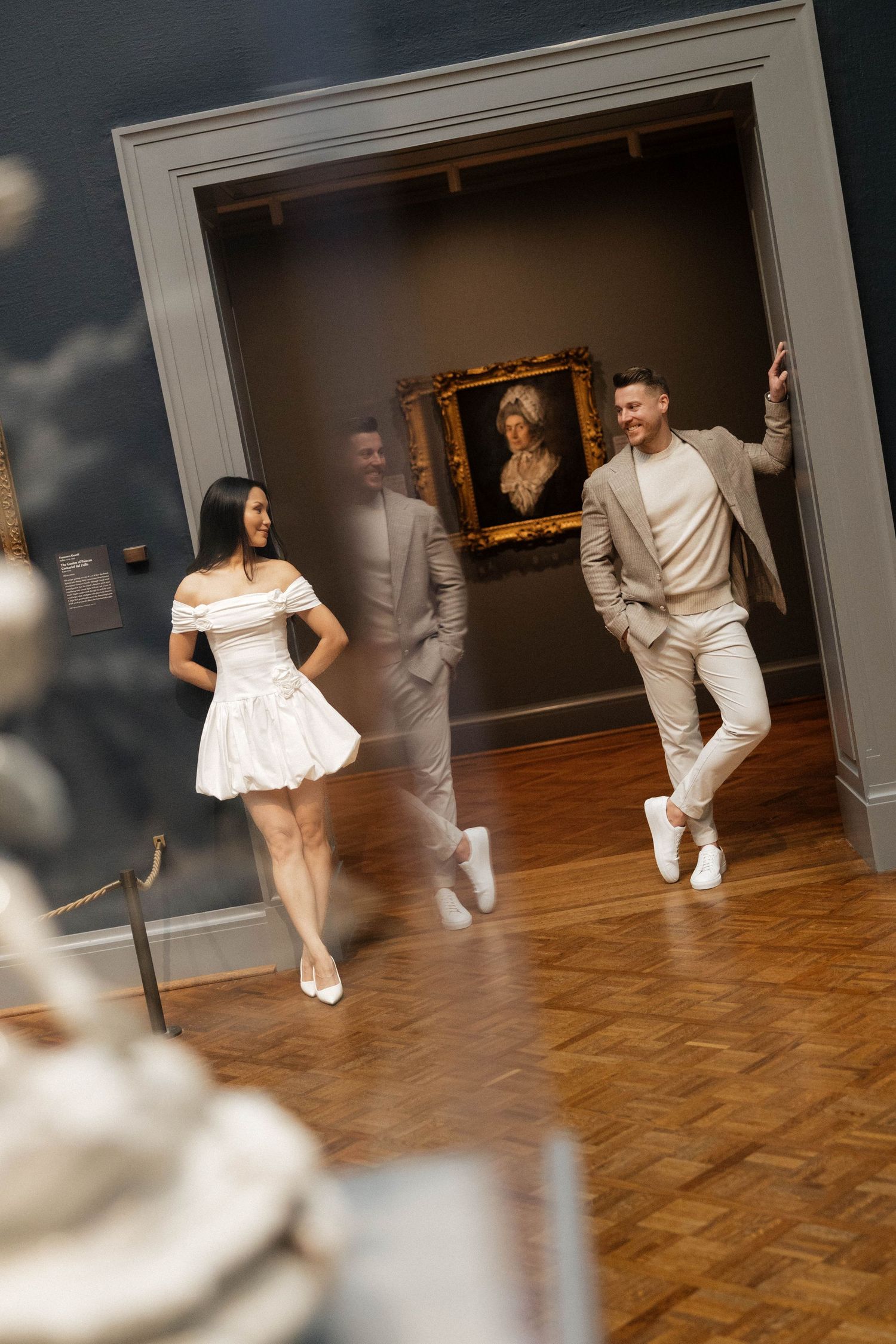 A stylish couple walks through an art museum gallery, with a classic portrait painting displayed on the wall behind them.