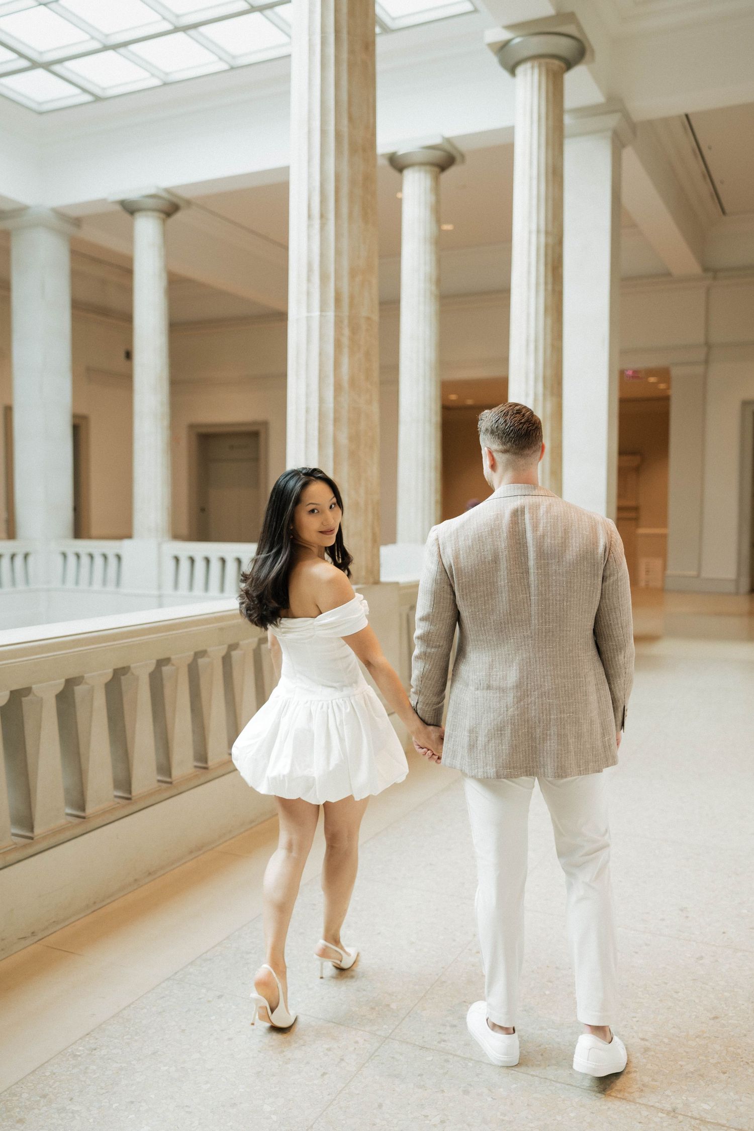 Couple holding hands walking through elegant museum hall with tall columns, woman in white mini dress, man in beige suit.