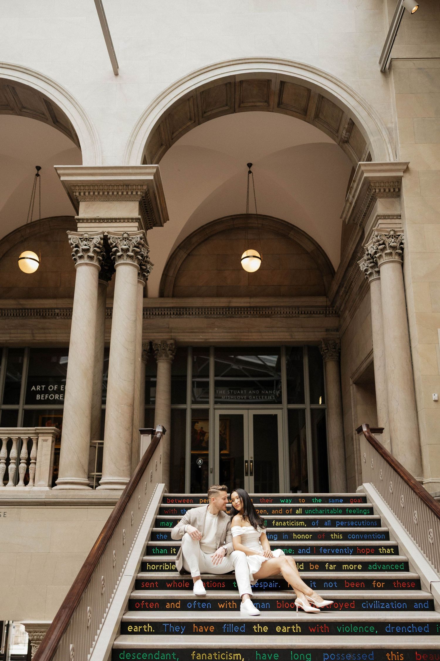 Couple sitting on colorful quote-inscribed steps of a grand neoclassical building with arched columns.