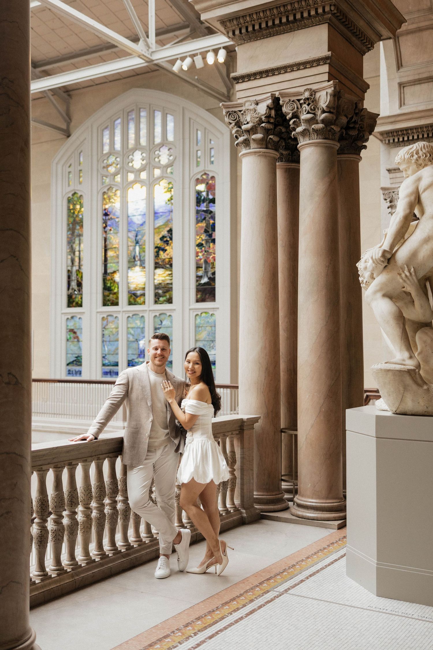 Couple posing in elegant museum hall with stained glass window, marble columns, and classical sculpture.