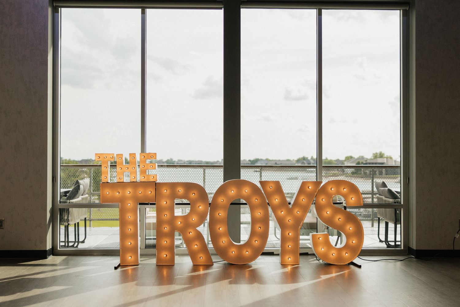Illuminated marquee letters spelling THE TROYS displayed in a bright venue with large windows overlooking water.