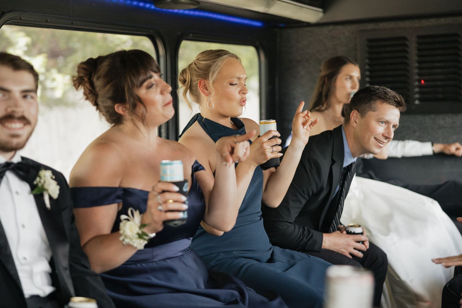 Wedding party guests in navy formal wear celebrate in a limousine, holding drinks and smiling during bridal party transportation.