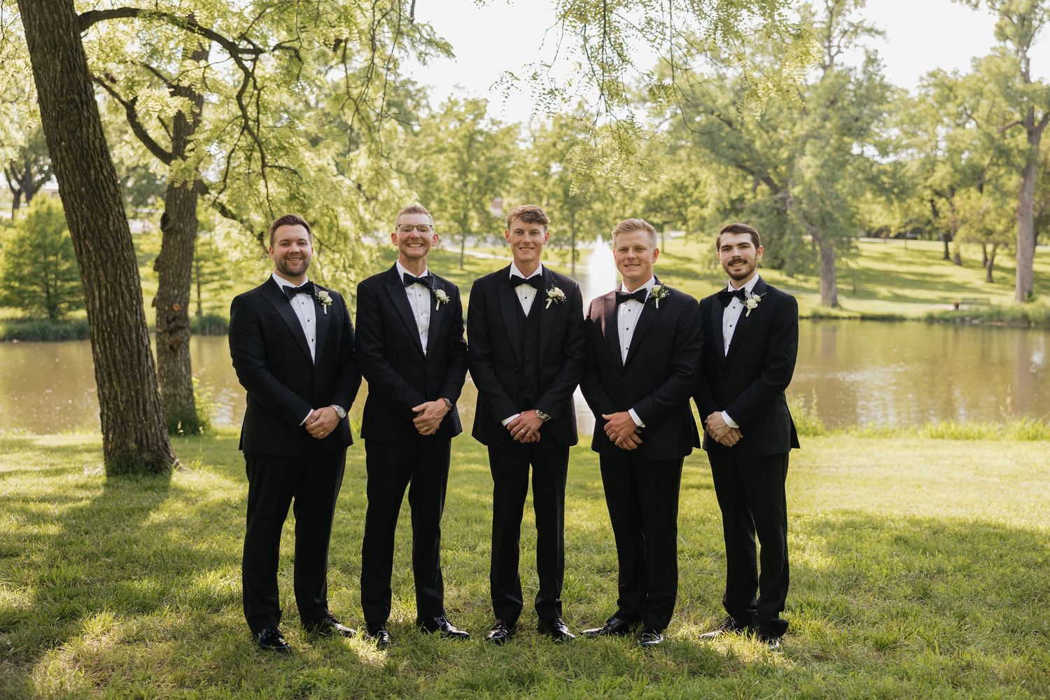 Five groomsmen in black tuxedos with bow ties posing outdoors near a pond surrounded by lush green trees.