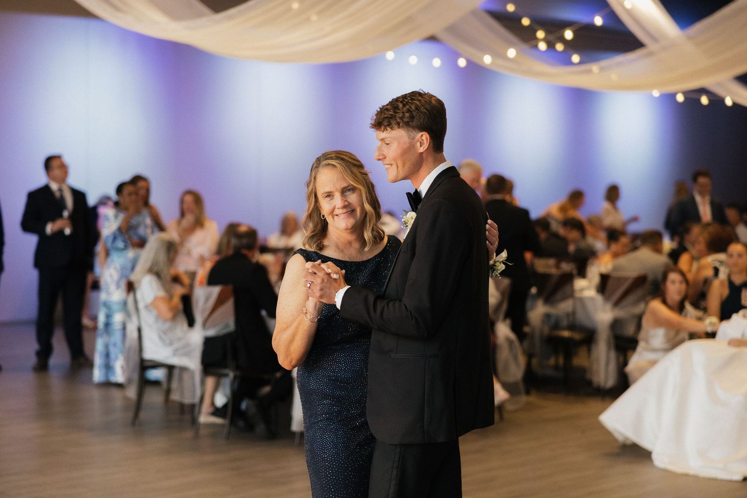 Mother and son share a tender dance at an elegant wedding reception with purple ambient lighting.