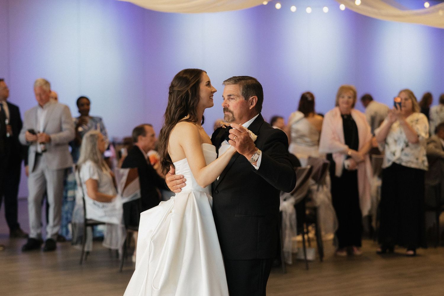 Bride and father share an emotional first dance at a wedding reception with guests watching in the background.