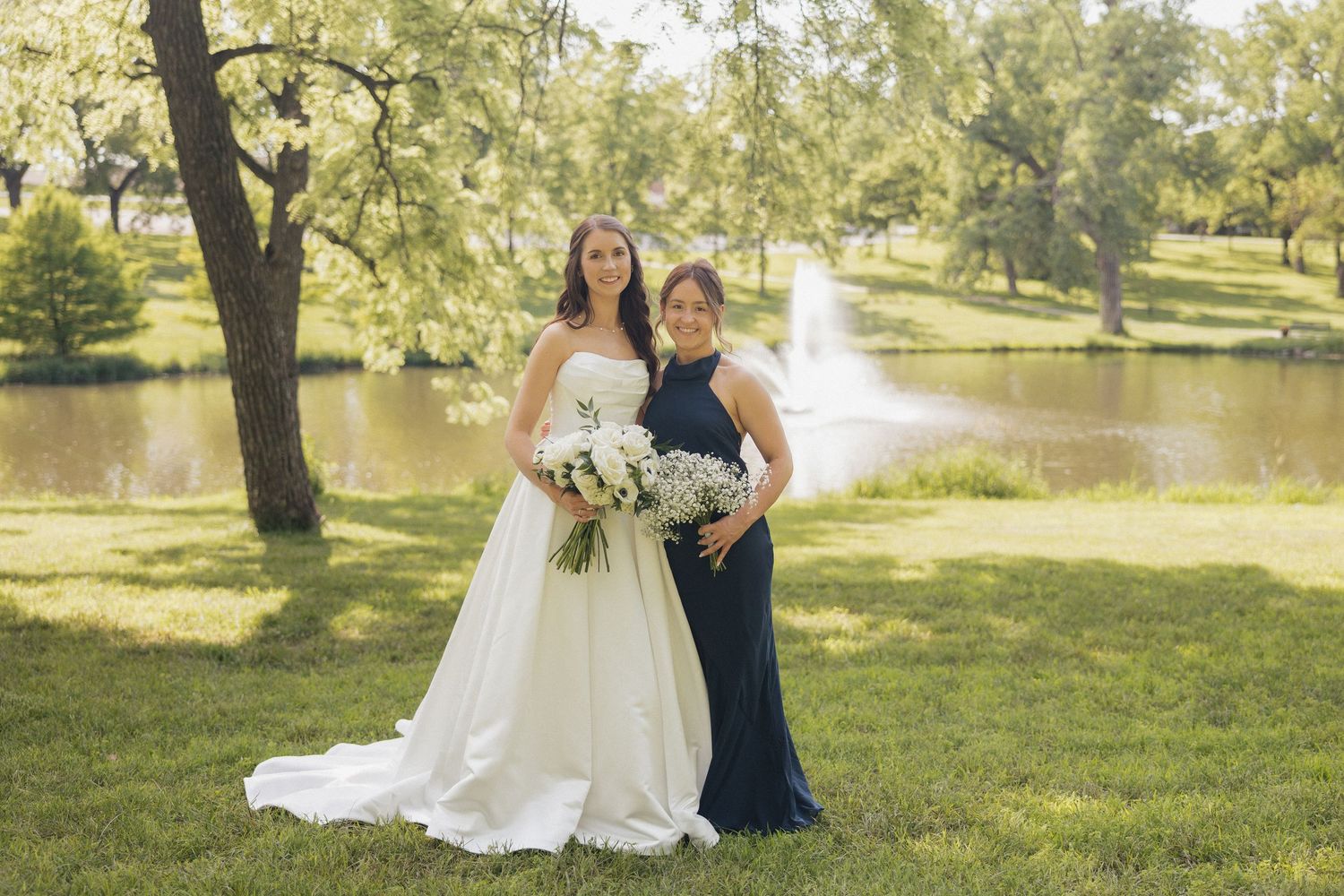 Bride in white gown and bridesmaid in navy dress pose by a pond with fountain in a lush green park.