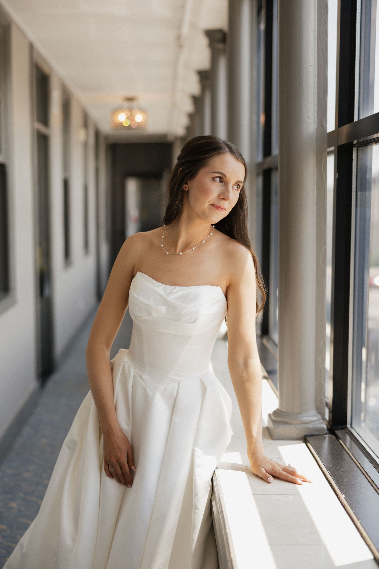 Bride in strapless white ball gown gazes out window in elegant hallway, wearing pearl necklace.