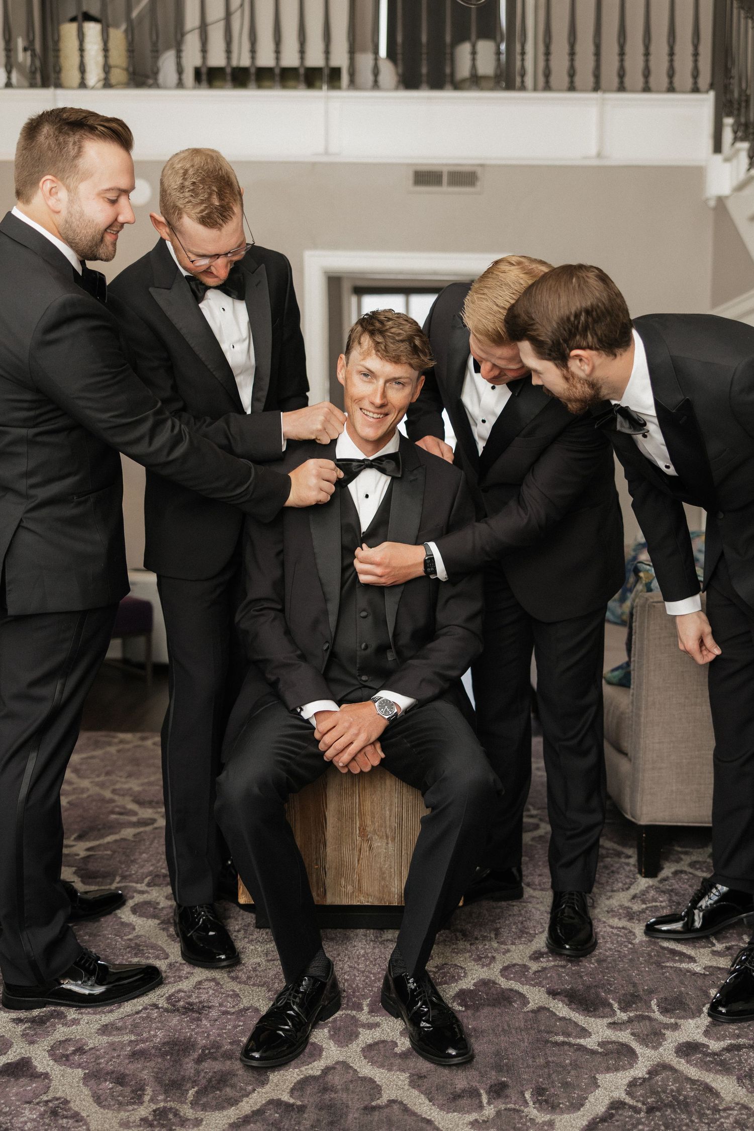 Groom sitting and smiling in black tuxedo while four groomsmen in matching suits help him get ready for wedding.