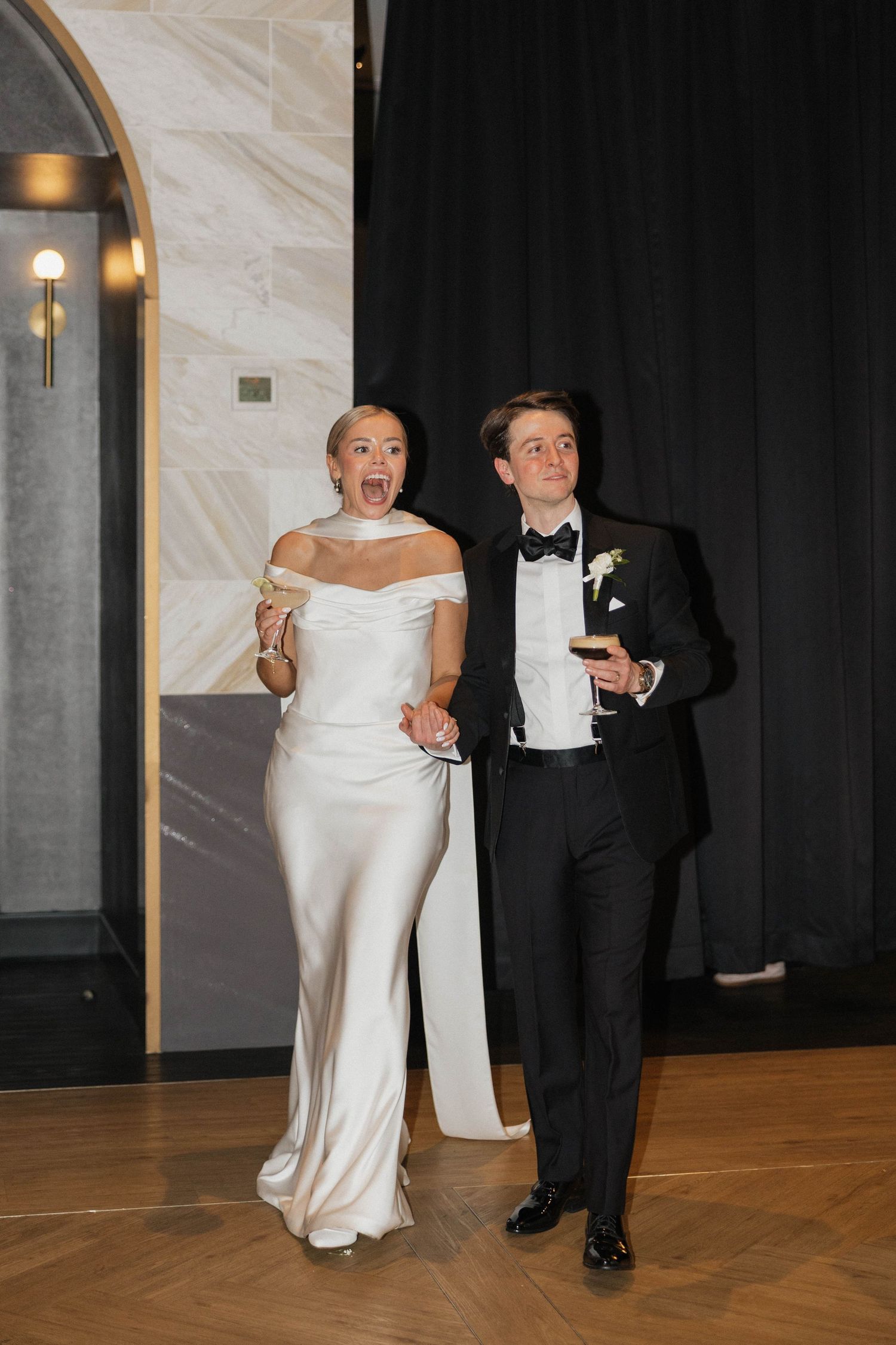 Bride in white satin gown and groom in black tuxedo making grand entrance at elegant wedding reception.