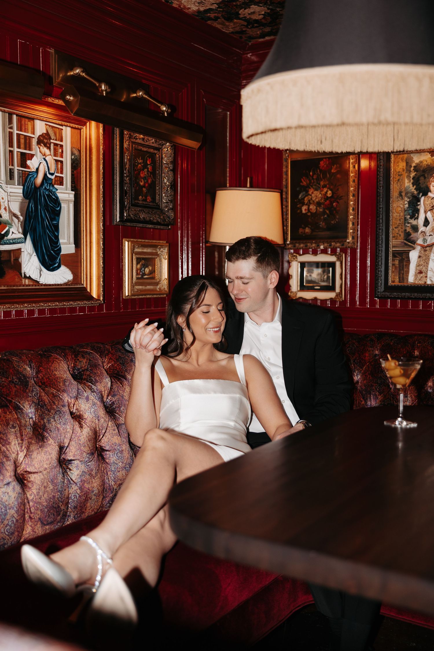 Couple relaxes in vintage bar with red walls, tufted leather seating and ornate decor illuminated by warm lighting.