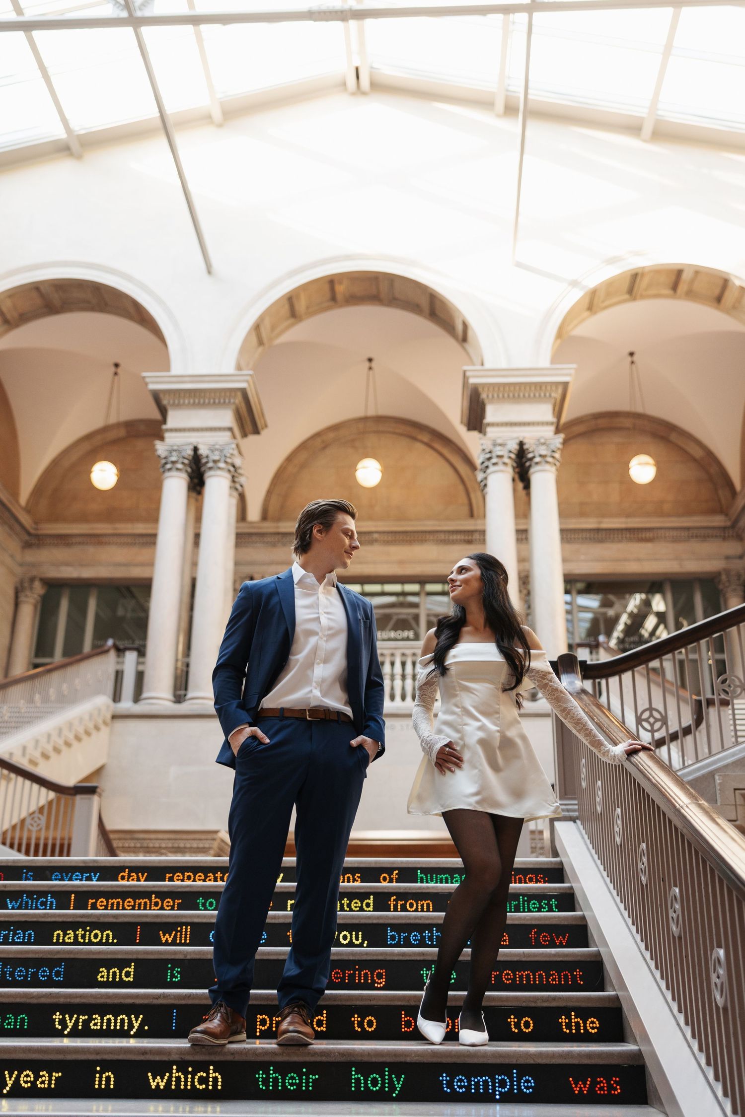 A stylish couple poses on architectural steps with elegant arched columns and natural lighting.