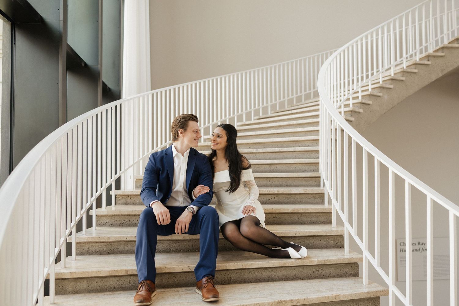 A couple sits intimately on elegant white stairs in formal attire.