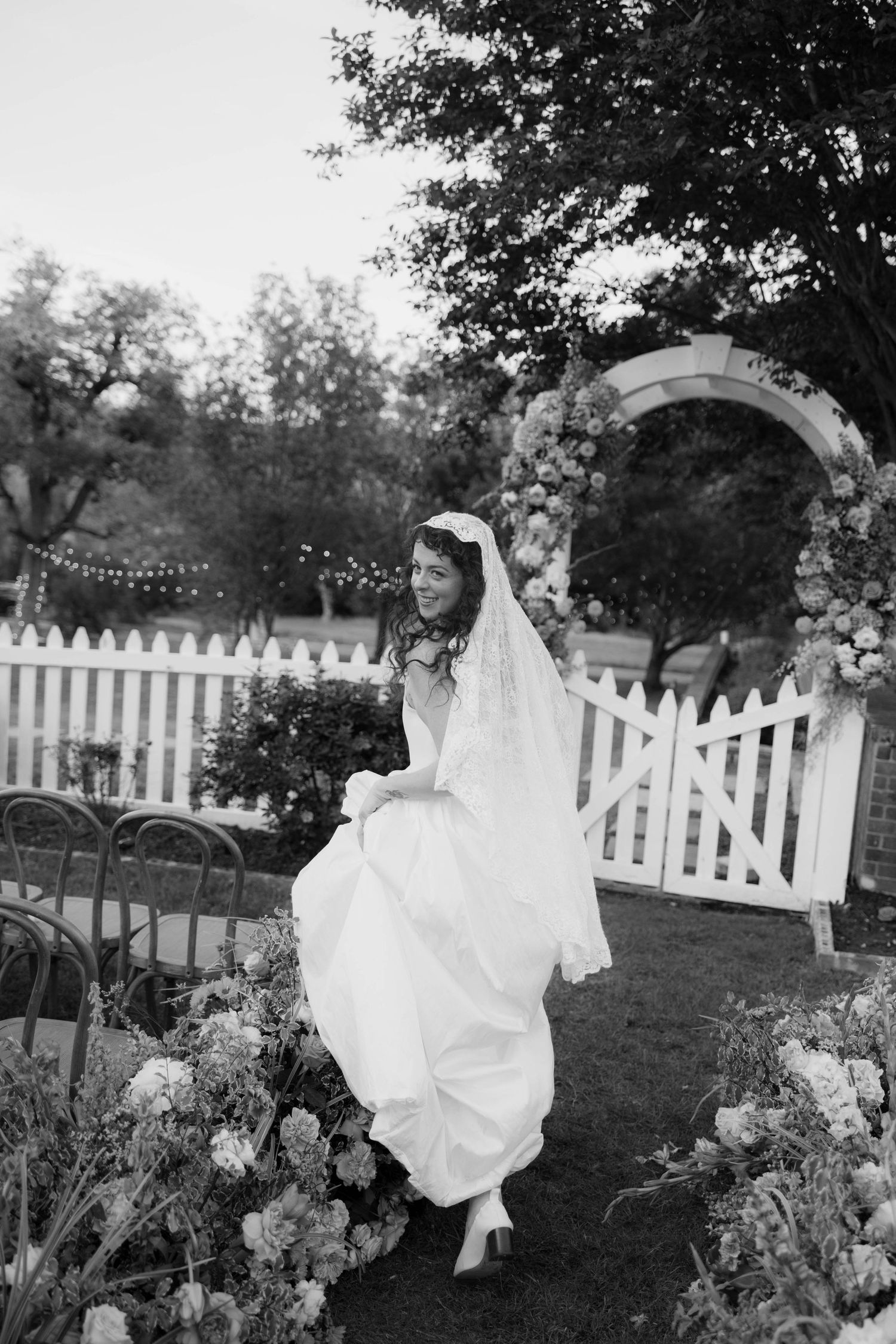Bride in white gown and veil walks through floral garden aisle near white picket fence and flower arch.