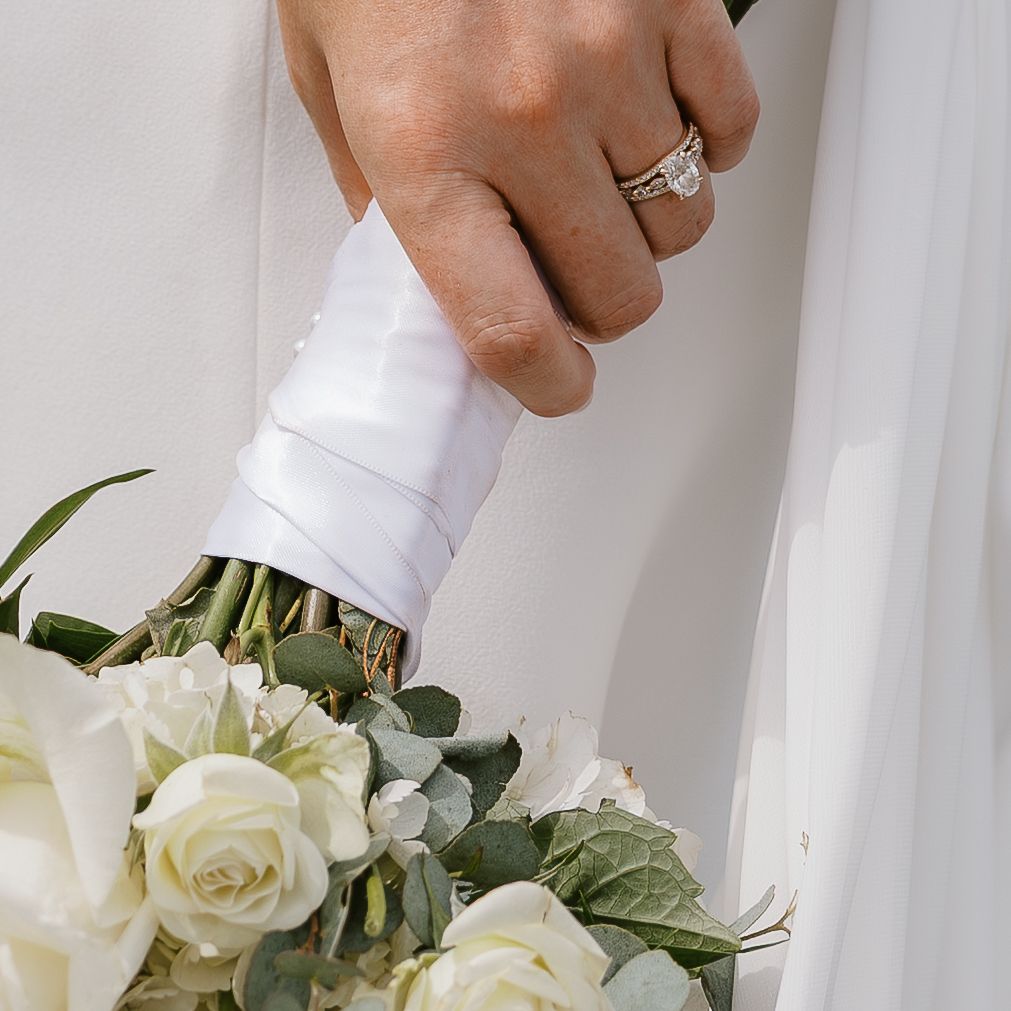 A person's hands delicately arranging white roses in a bouquet with green foliage against a light background.