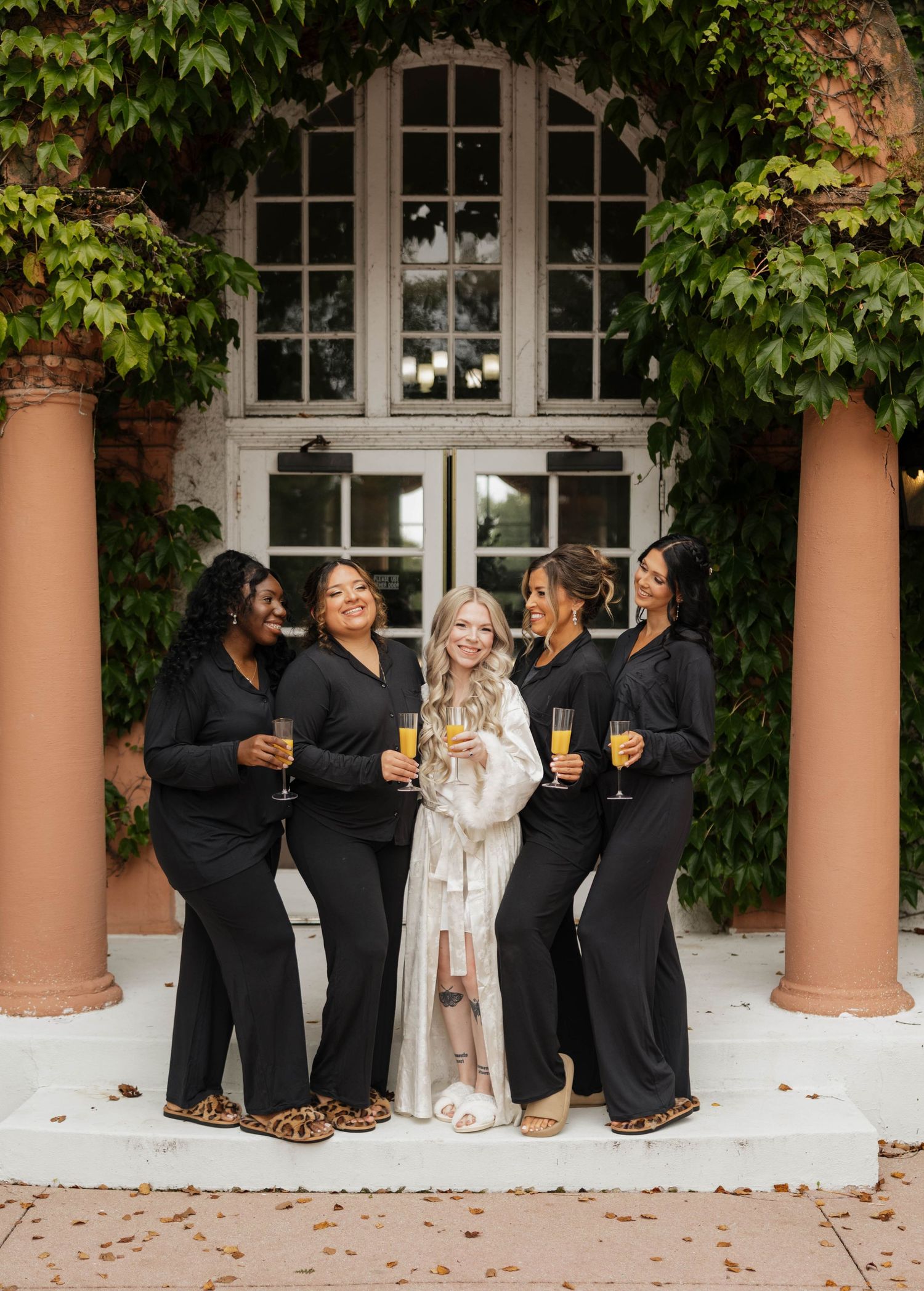 Five women in black loungewear and a bride in a white robe hold champagne glasses on stone steps outside a vine-covered brick building.