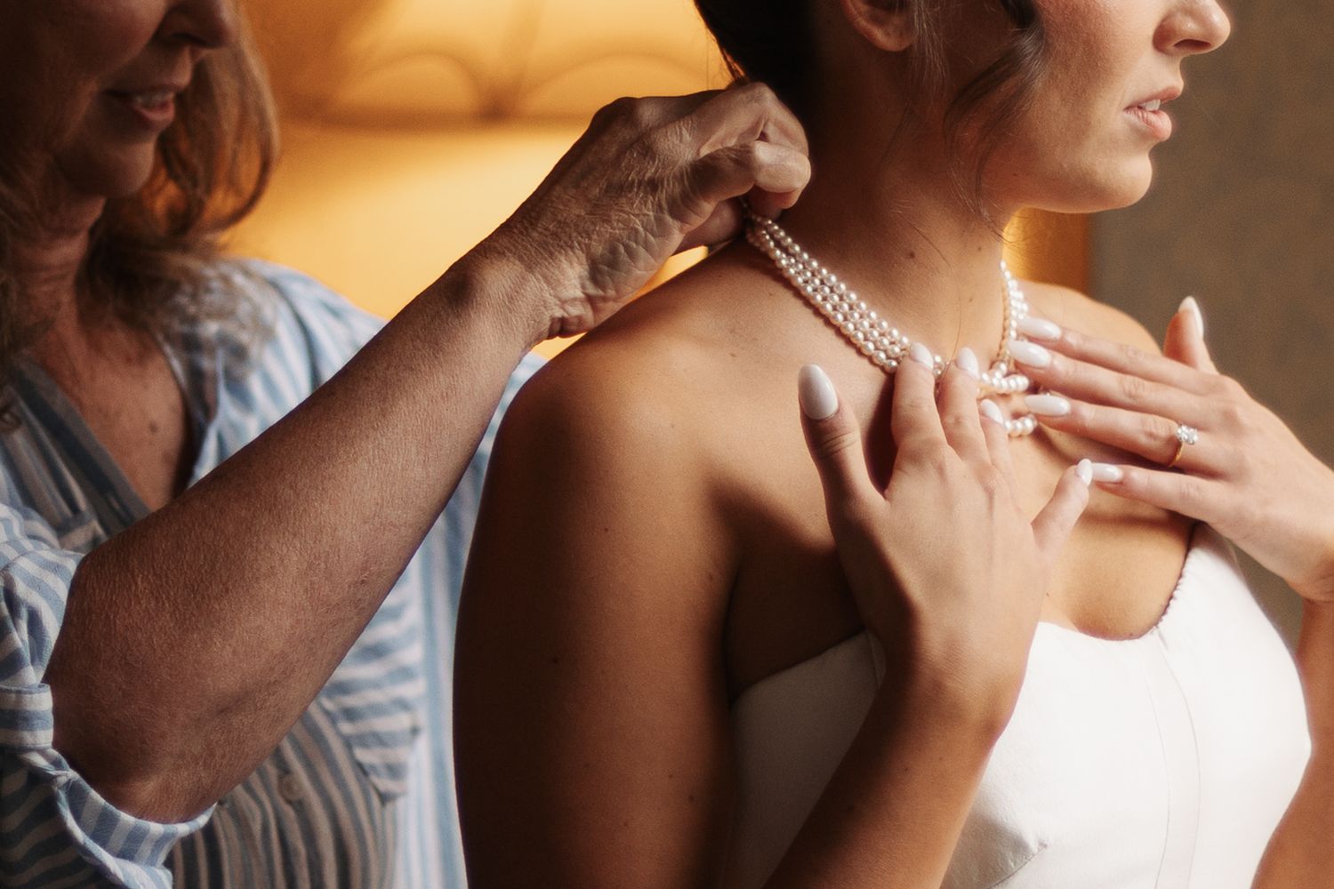 Older woman's hands fastening a pearl necklace around the neck of a bride in a white dress, a tender wedding day moment.