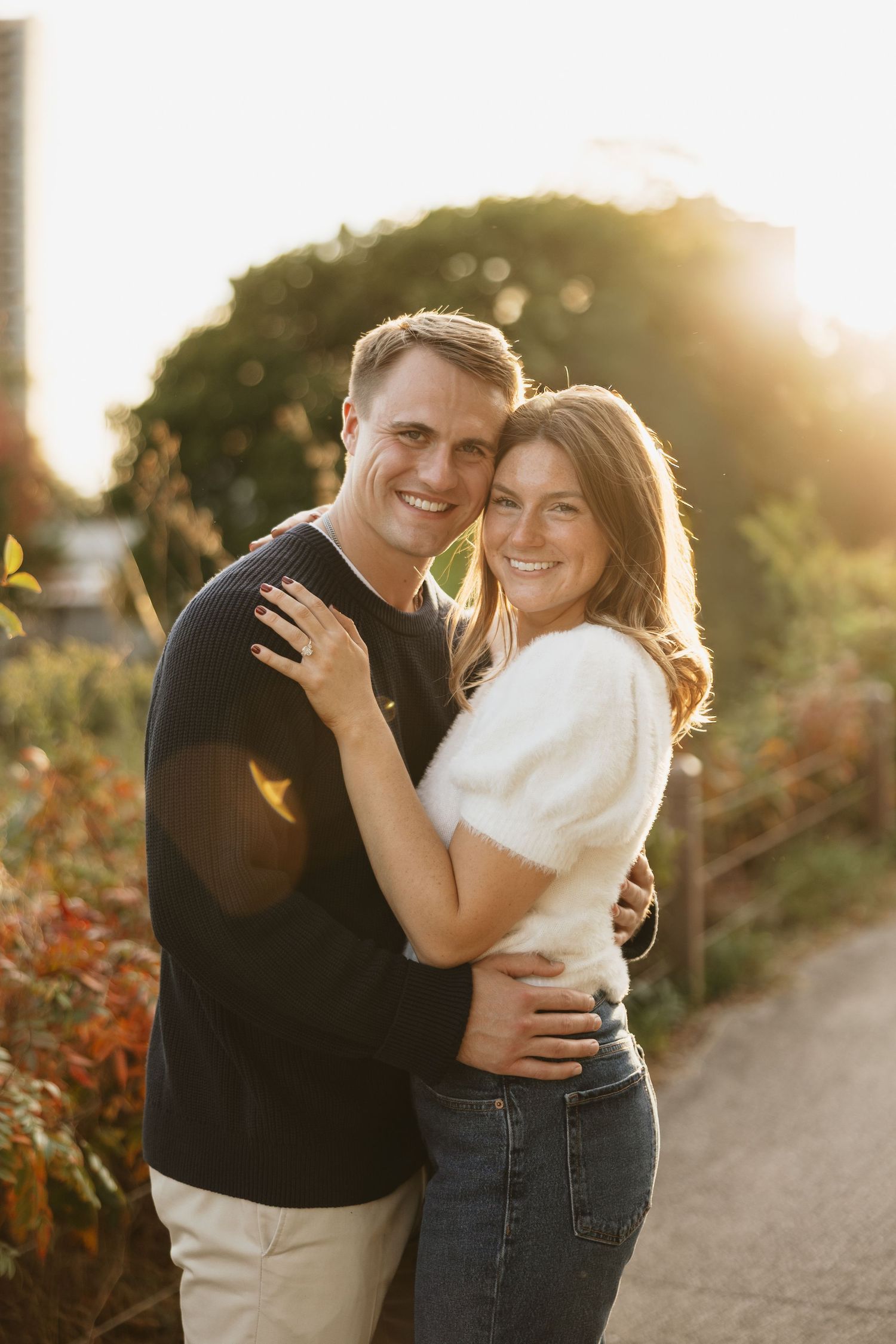 Smiling couple embracing outdoors at golden hour, the woman wearing a white sweater and the man a navy sweater, surrounded by greenery.