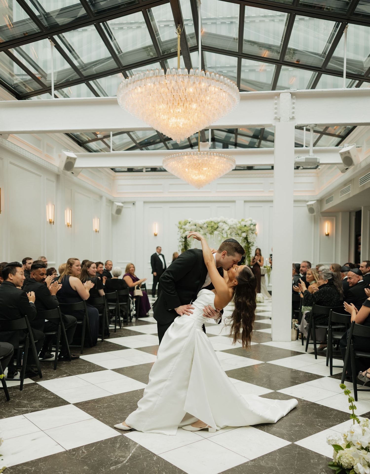 Bride and groom share a dramatic dip kiss on a black and white checkered dance floor under crystal chandeliers as wedding guests watch.