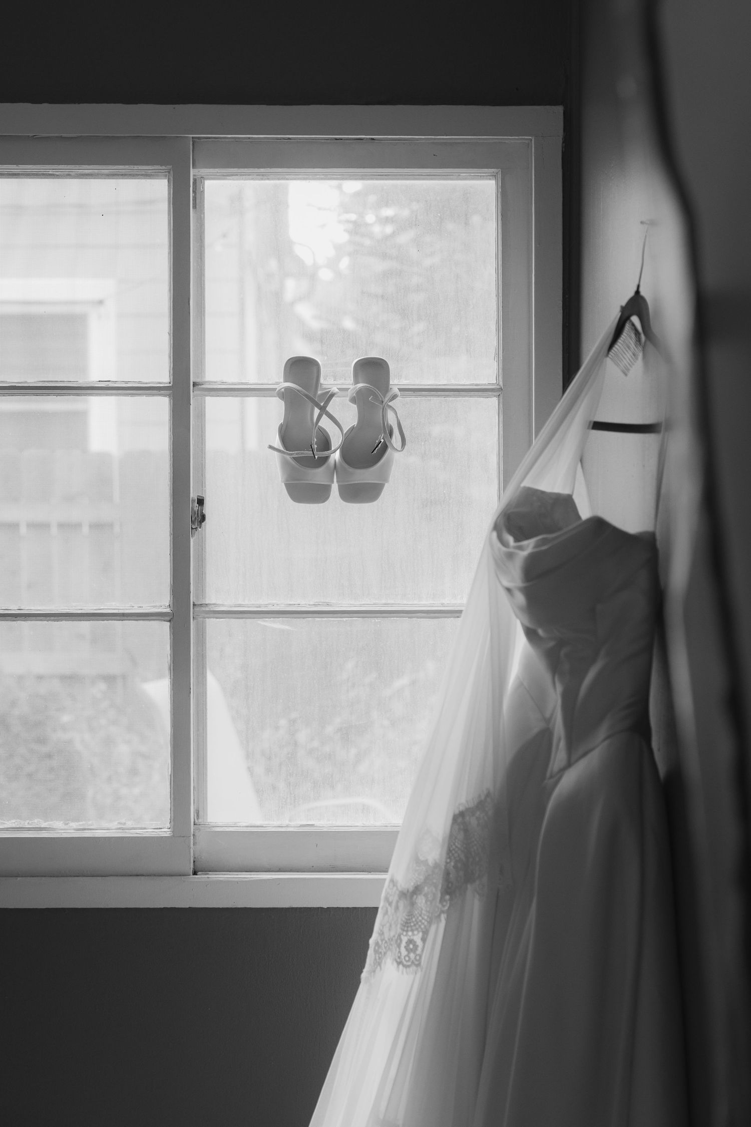 Black and white photo of a wedding dress with veil hanging beside a sunlit window with bridal sandals suspended in front of the glass.