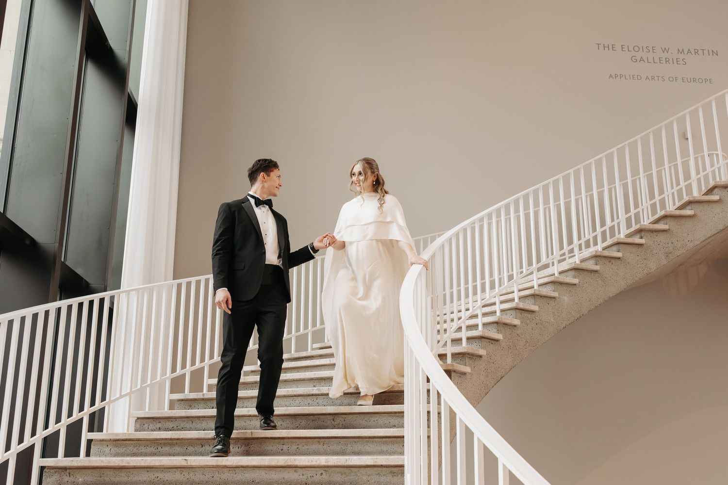 Bride and groom holding hands on elegant staircase in white wedding dress and black tuxedo at art gallery venue.