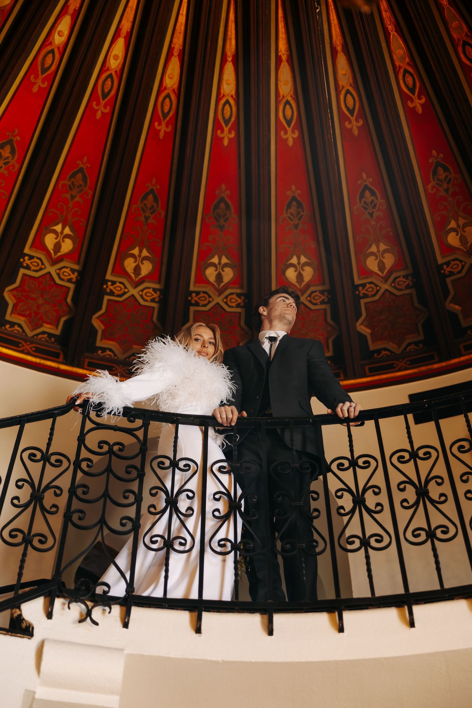 Wedding party stands along curved black iron railing with red and gold decorative panels in the background.