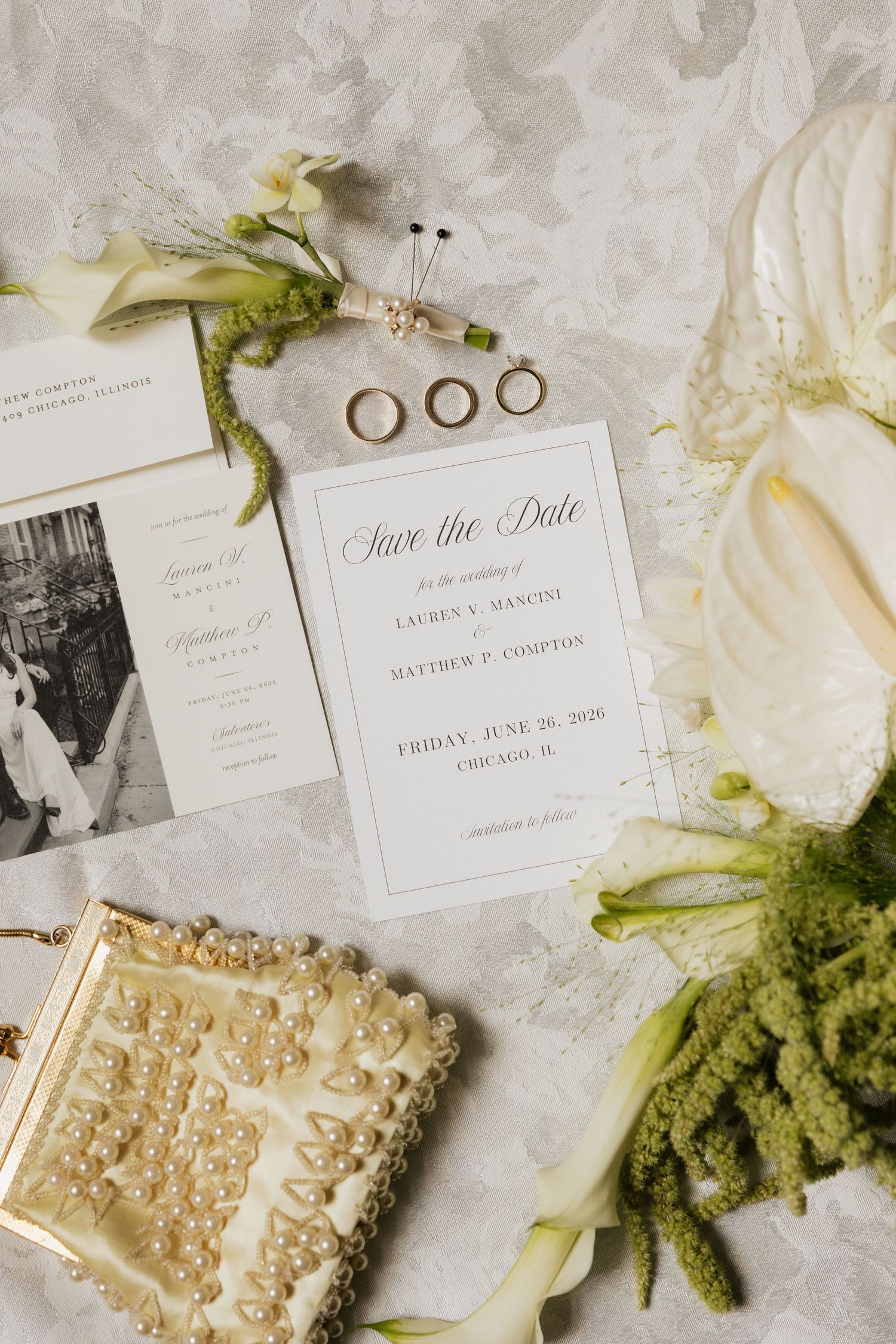 Elegant wedding flat lay with save the date cards, gold rings, pearl clutch, and white floral arrangements on marble.