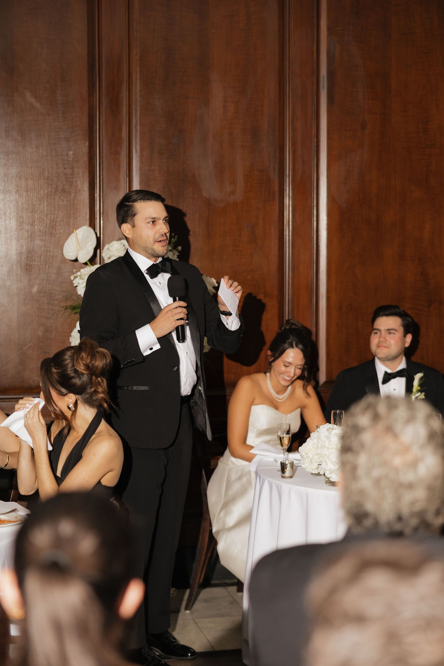 A man in a tuxedo gives a wedding toast while the bride smiles at a decorated reception table.