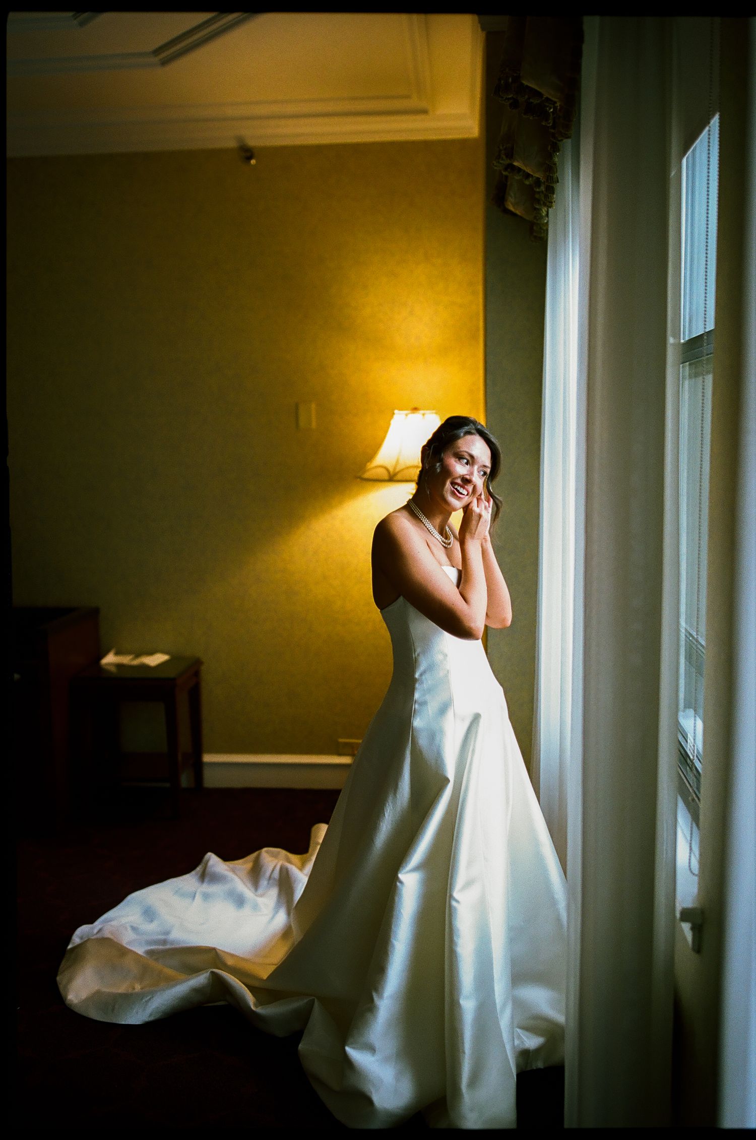Bride in elegant white wedding gown standing by a sunlit window, smiling and touching her face in a warmly lit room.