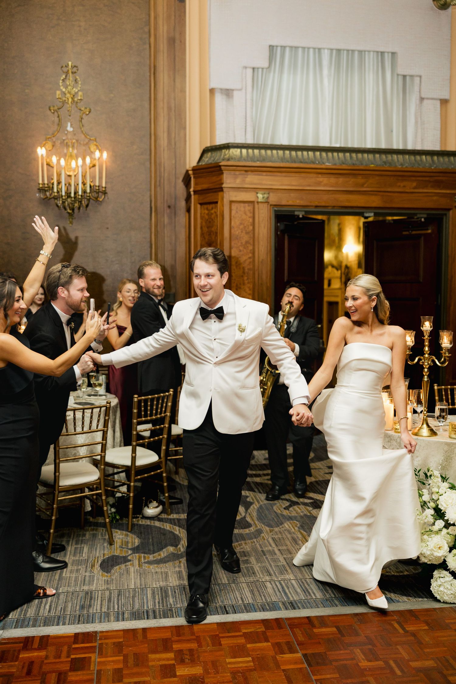 Smiling groom in white dinner jacket and bride in strapless gown enter their wedding reception holding hands as guests cheer.