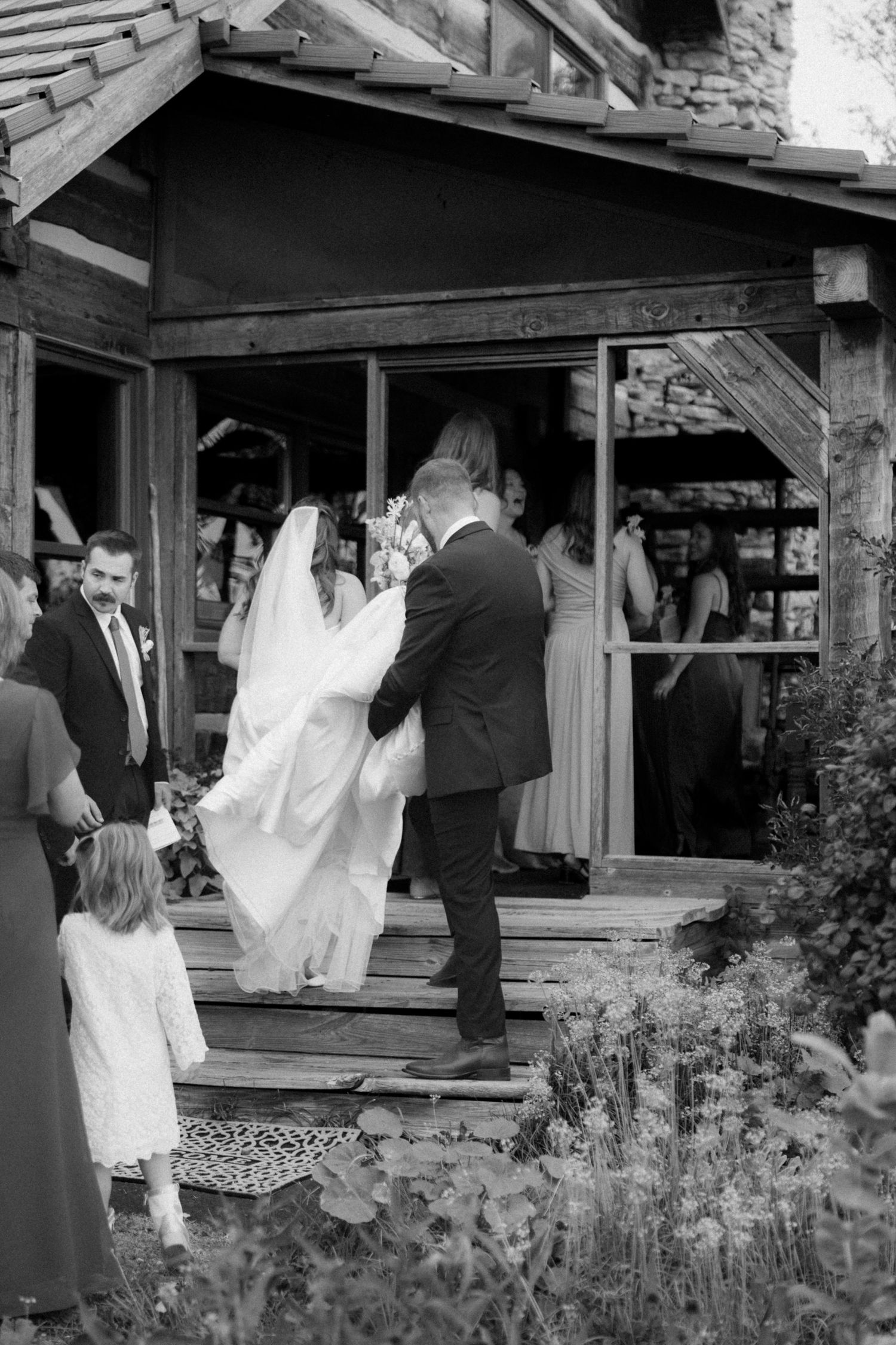 Black and white photo of a groom helping the bride up wooden steps as a flower girl and guests follow into a rustic venue.