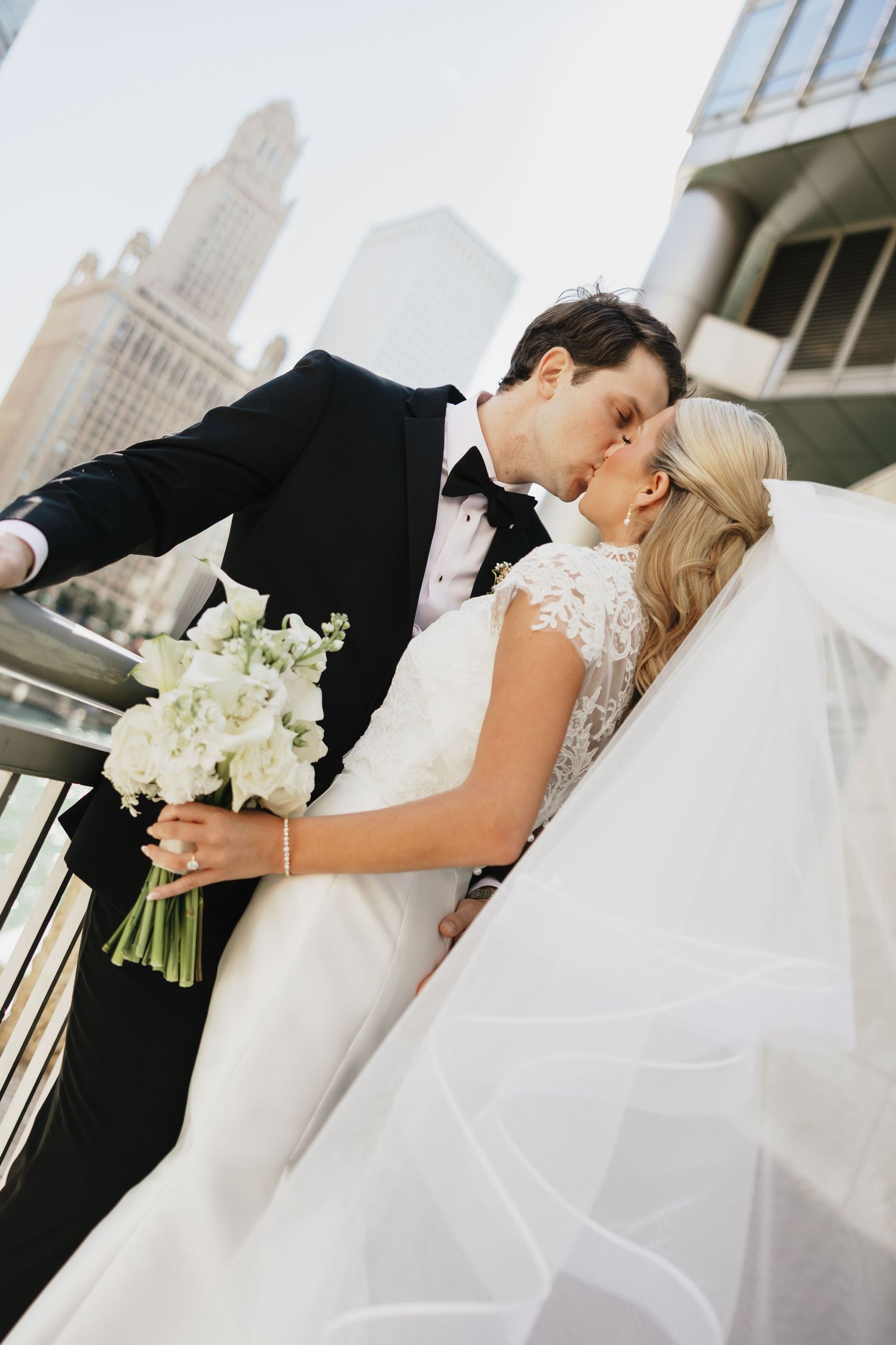 Bride in a lace-sleeved gown and long veil kisses a groom in a black tuxedo, holding a white floral bouquet on a city balcony.