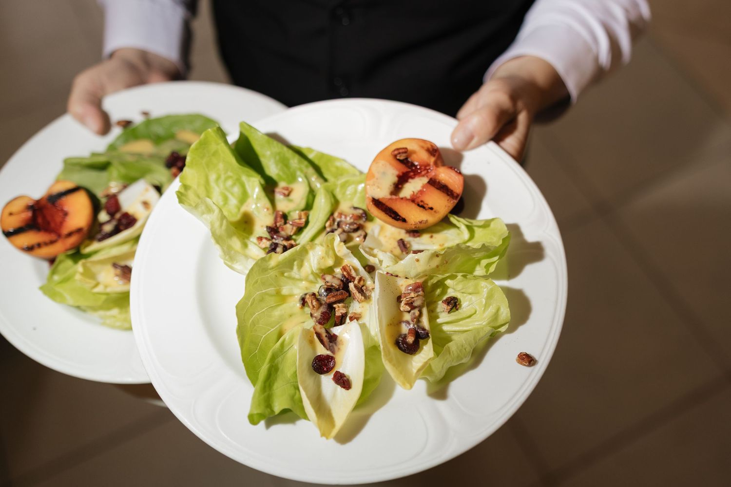 Waiter serving two white plates of fresh green salad with grilled peaches and nuts in a restaurant.