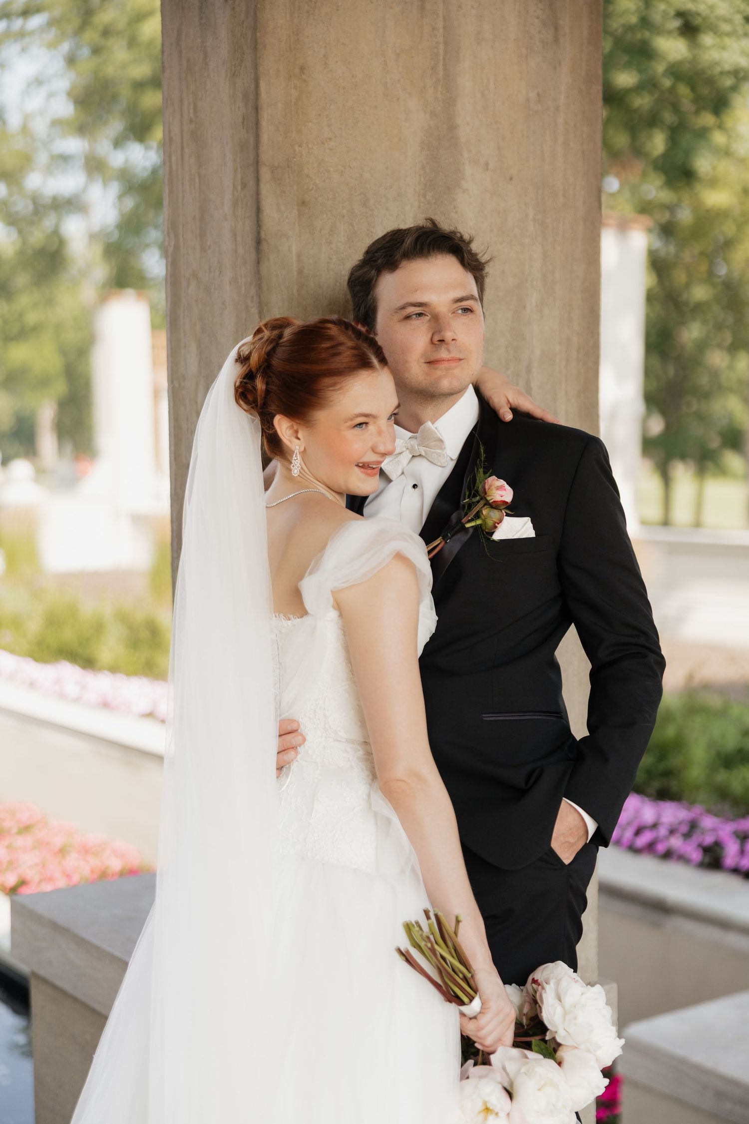 Bride in white gown with veil holds bouquet while groom in black suit embraces her by stone column.
