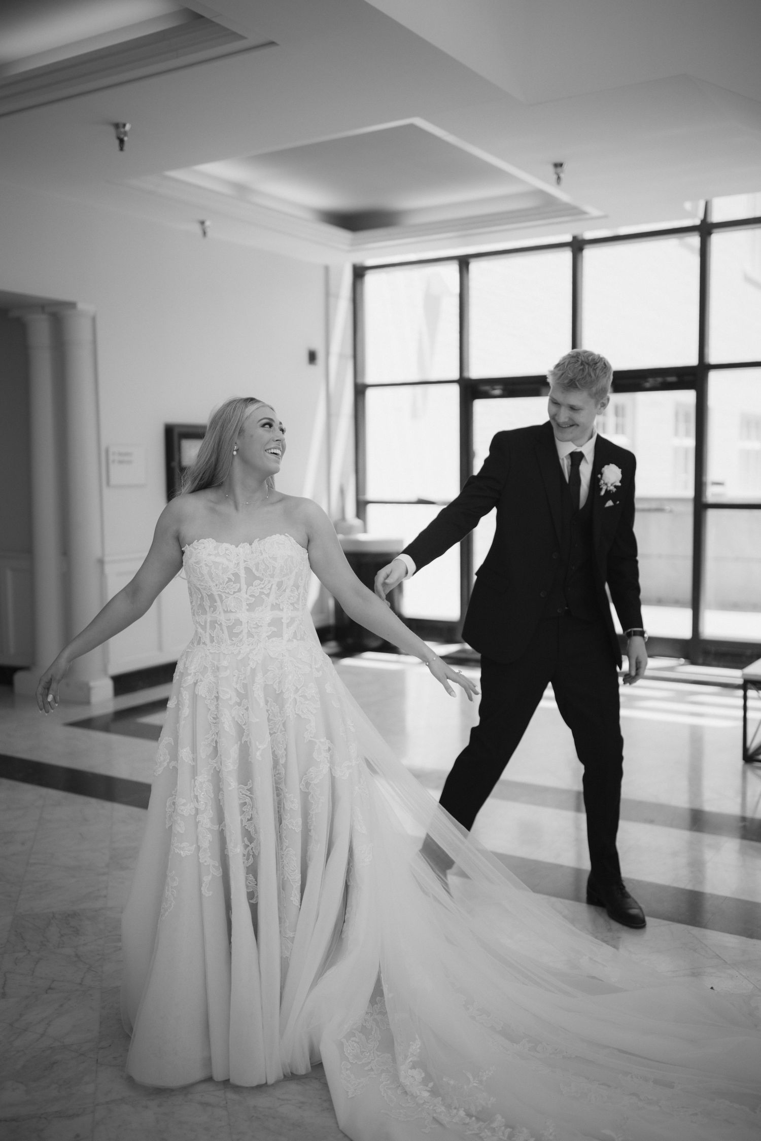 A bride in a flowing white gown dances with someone in formal wear across a bright open space.