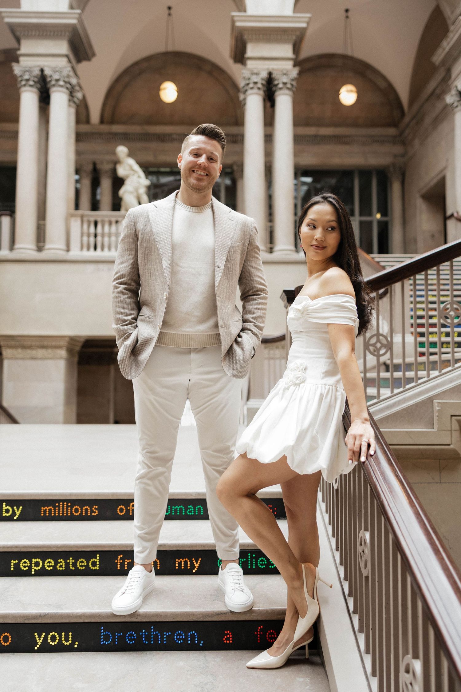 Elegant couple posing on ornate staircase, man in beige suit, woman in white off-shoulder mini dress, grand architecture.