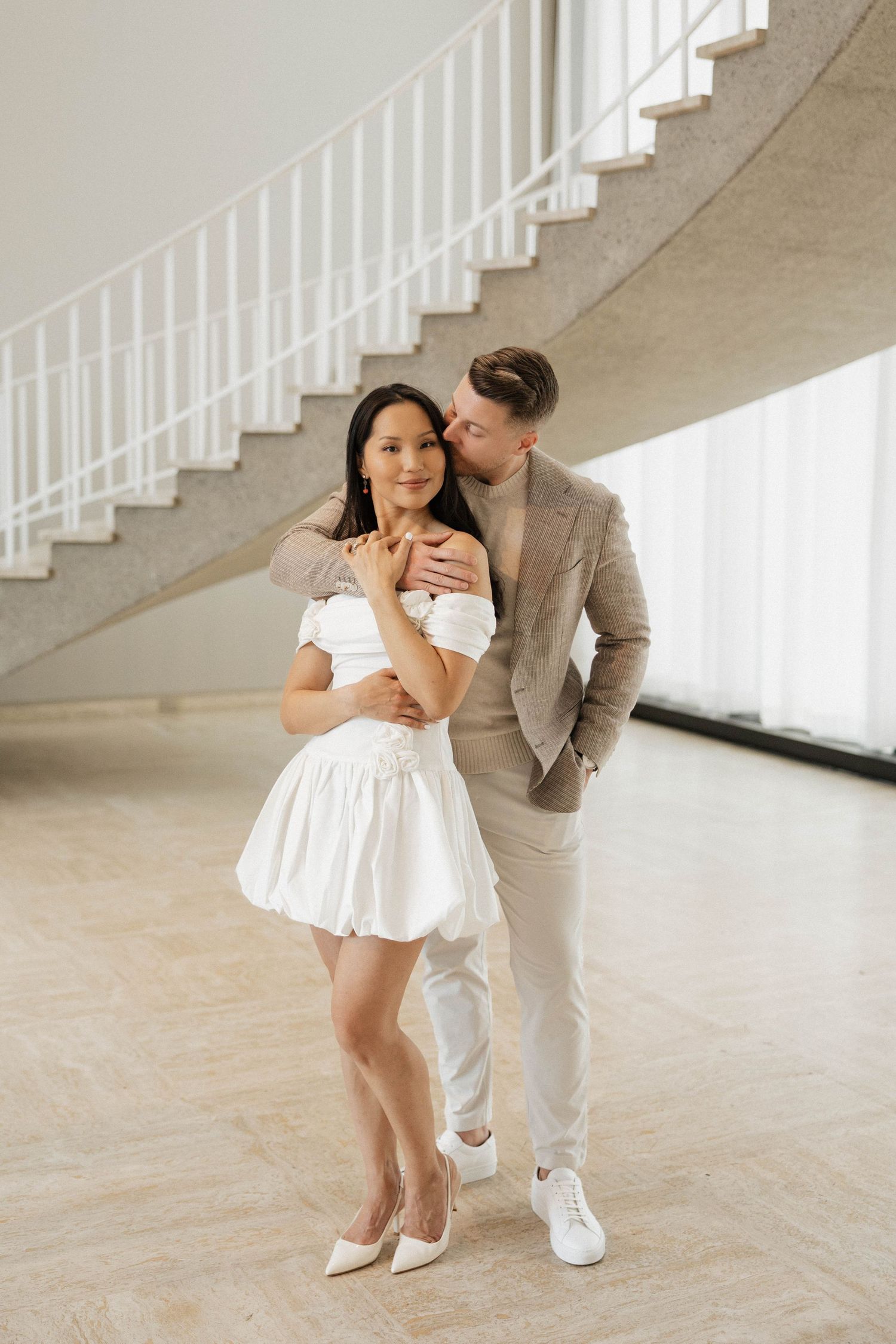 Couple posing by curved staircase, man in beige blazer embracing woman in white off-shoulder mini dress.