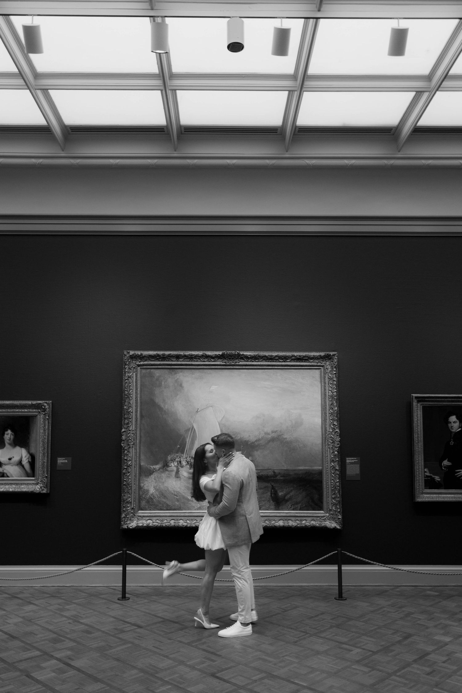 Black and white photo of a couple embracing in a museum gallery in front of a large classical painting.