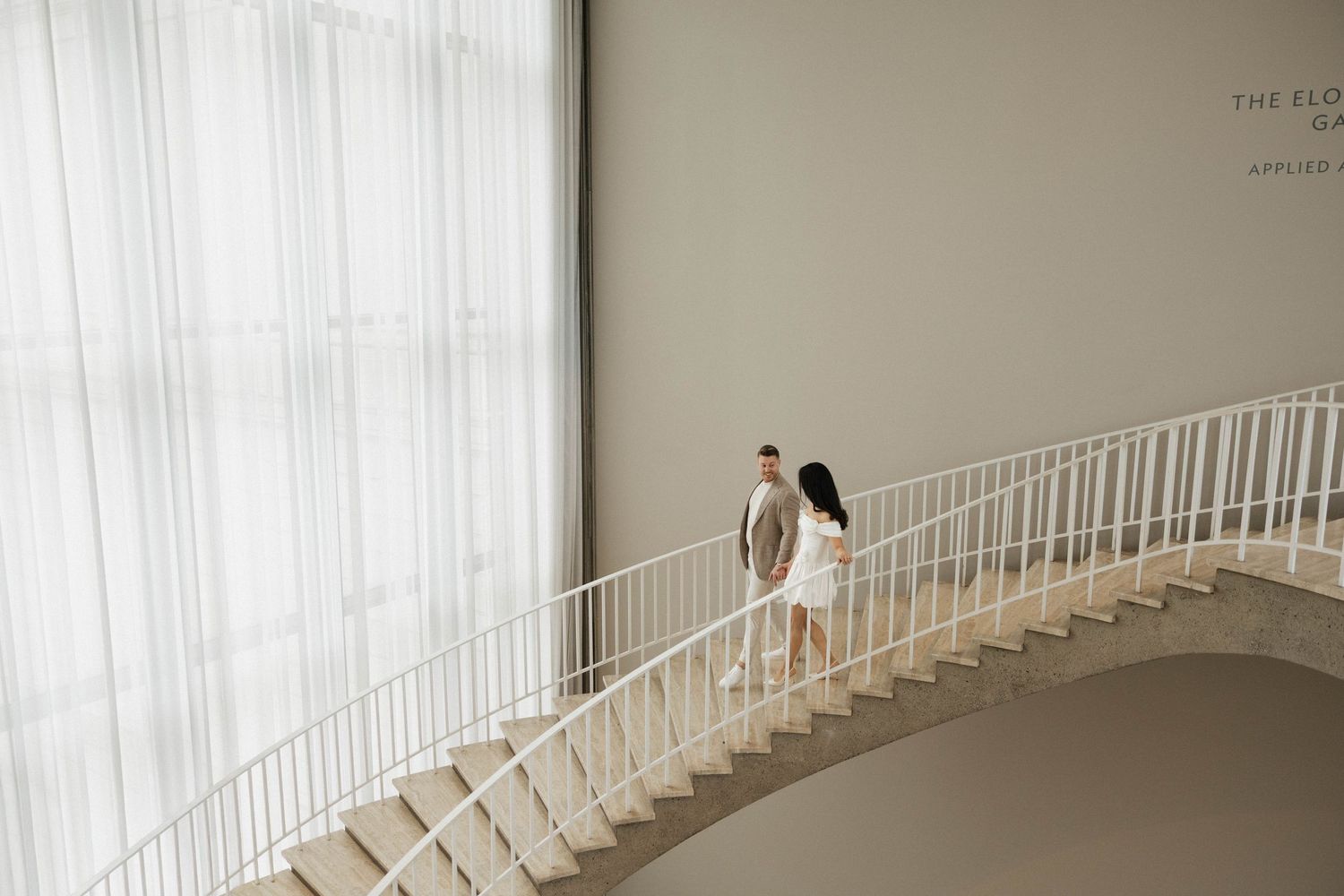 Couple holding hands walking up elegant curved white staircase in modern minimalist interior with sheer curtains.