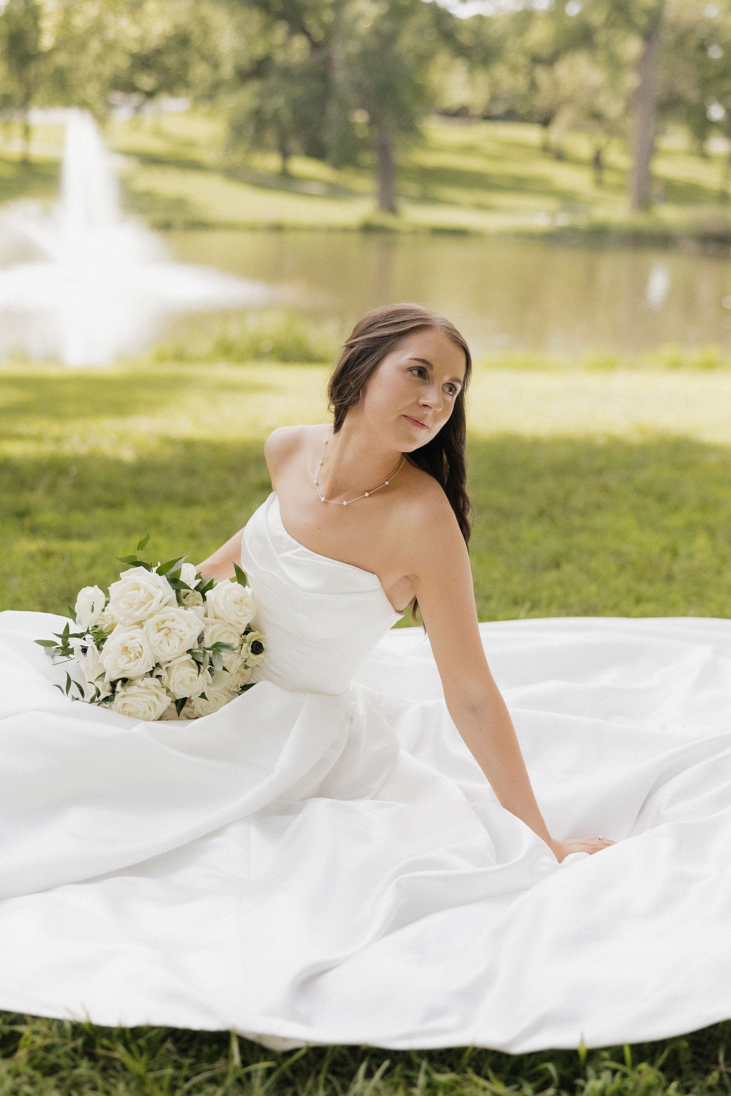 Bride in strapless white ball gown sitting on grass holding white rose bouquet with pond and fountain backdrop.