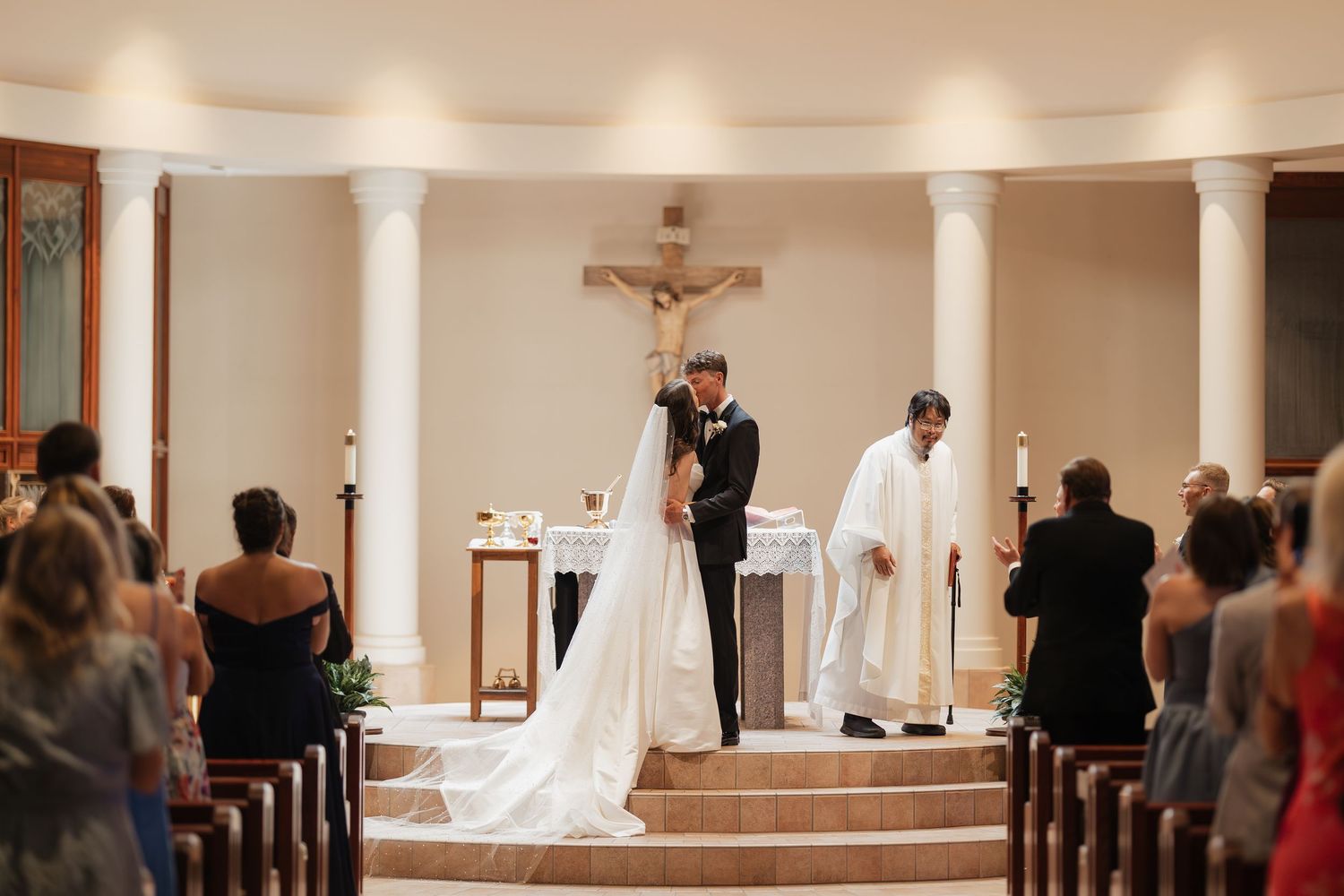 Bride and groom share first kiss at church altar during wedding ceremony with officiant and guests watching.