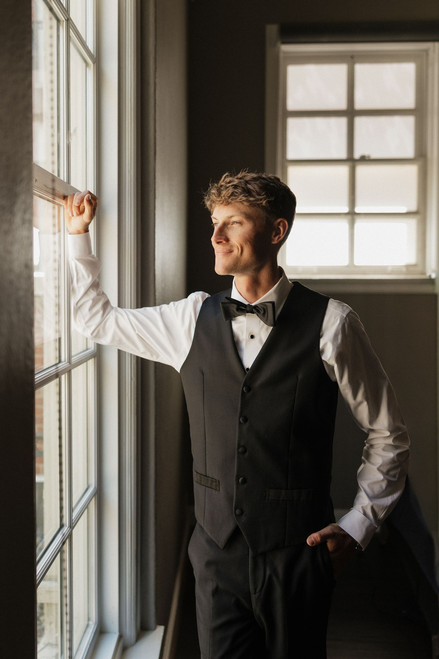 Elegant groom in black vest and bow tie gazing out a sunlit window on his wedding day.