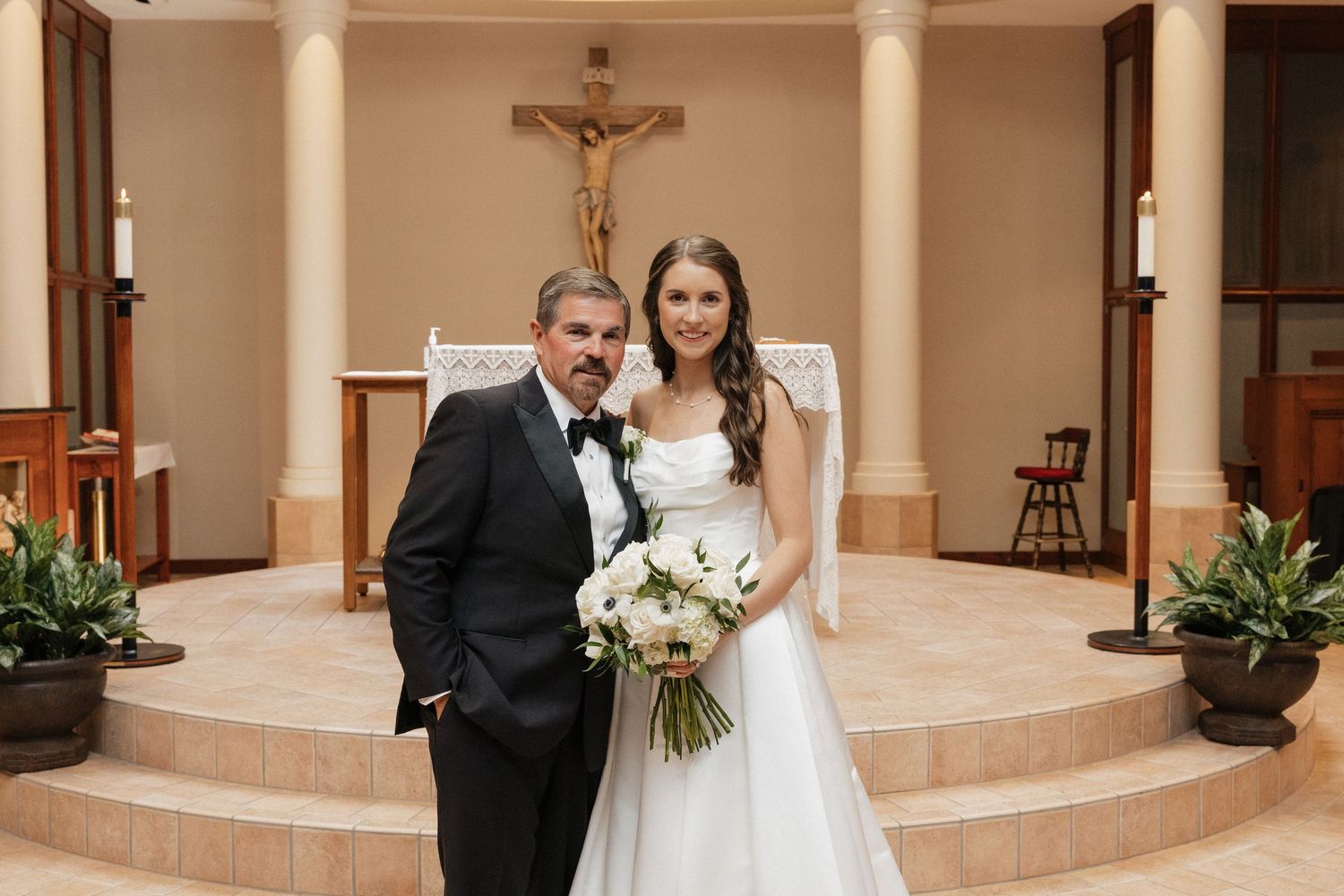 Bride in white gown holding bouquet stands with man in tuxedo before altar in elegant chapel.