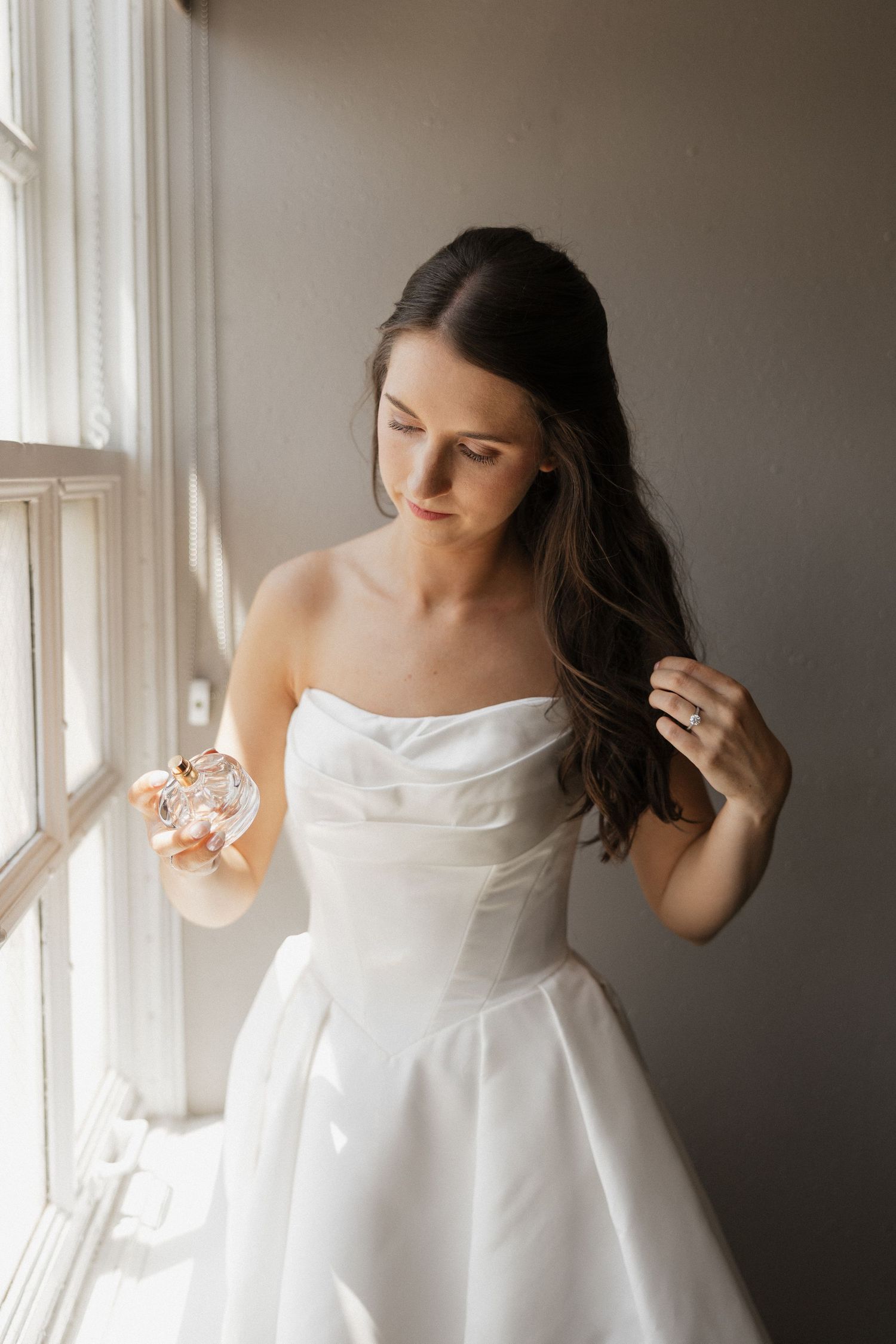 Bride in strapless white gown holding perfume bottle by window, dark hair flowing, wearing engagement ring.