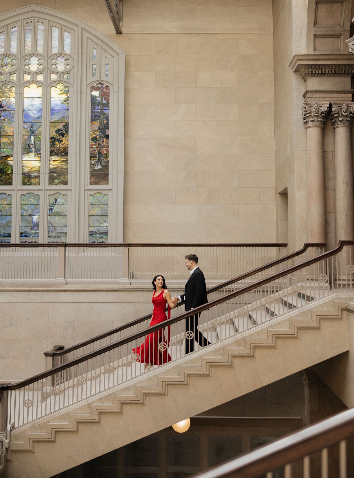 A couple descends an elegant staircase in formal attire against a backdrop of stained glass windows.