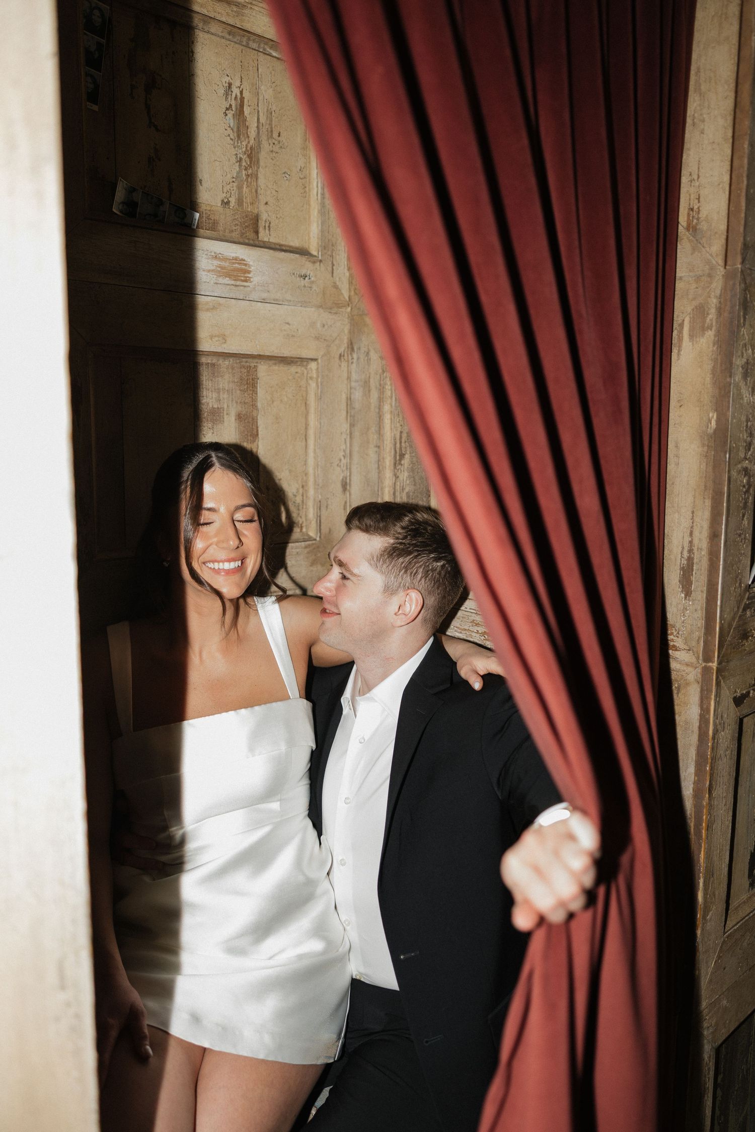 A couple in formal attire stands near red curtains in a dimly lit venue during a celebratory event.