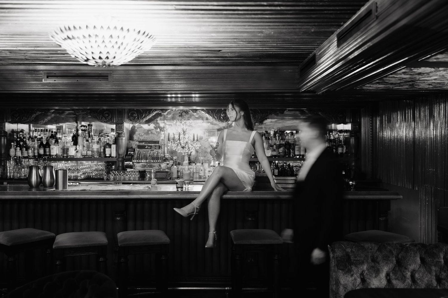 Black and white photo of an elegant bar interior with crystal chandelier and someone perched on the counter.