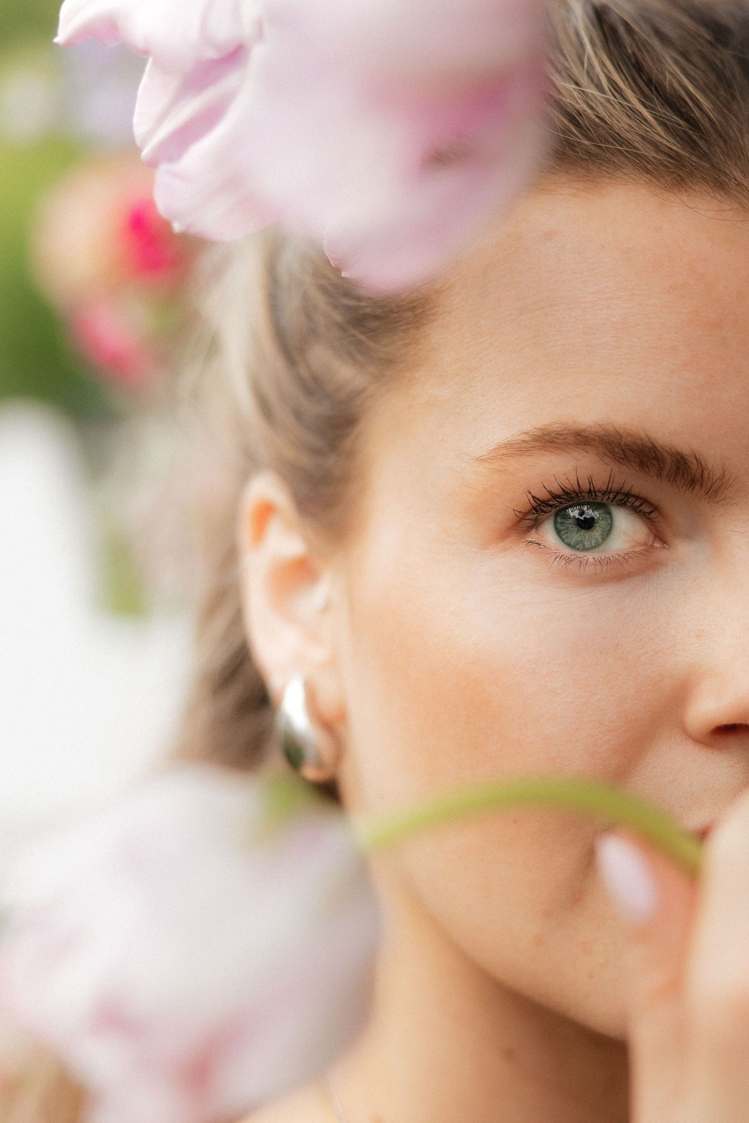 Close-up portrait of a young woman with blue-green eyes and silver hoop earring surrounded by soft pink flowers.