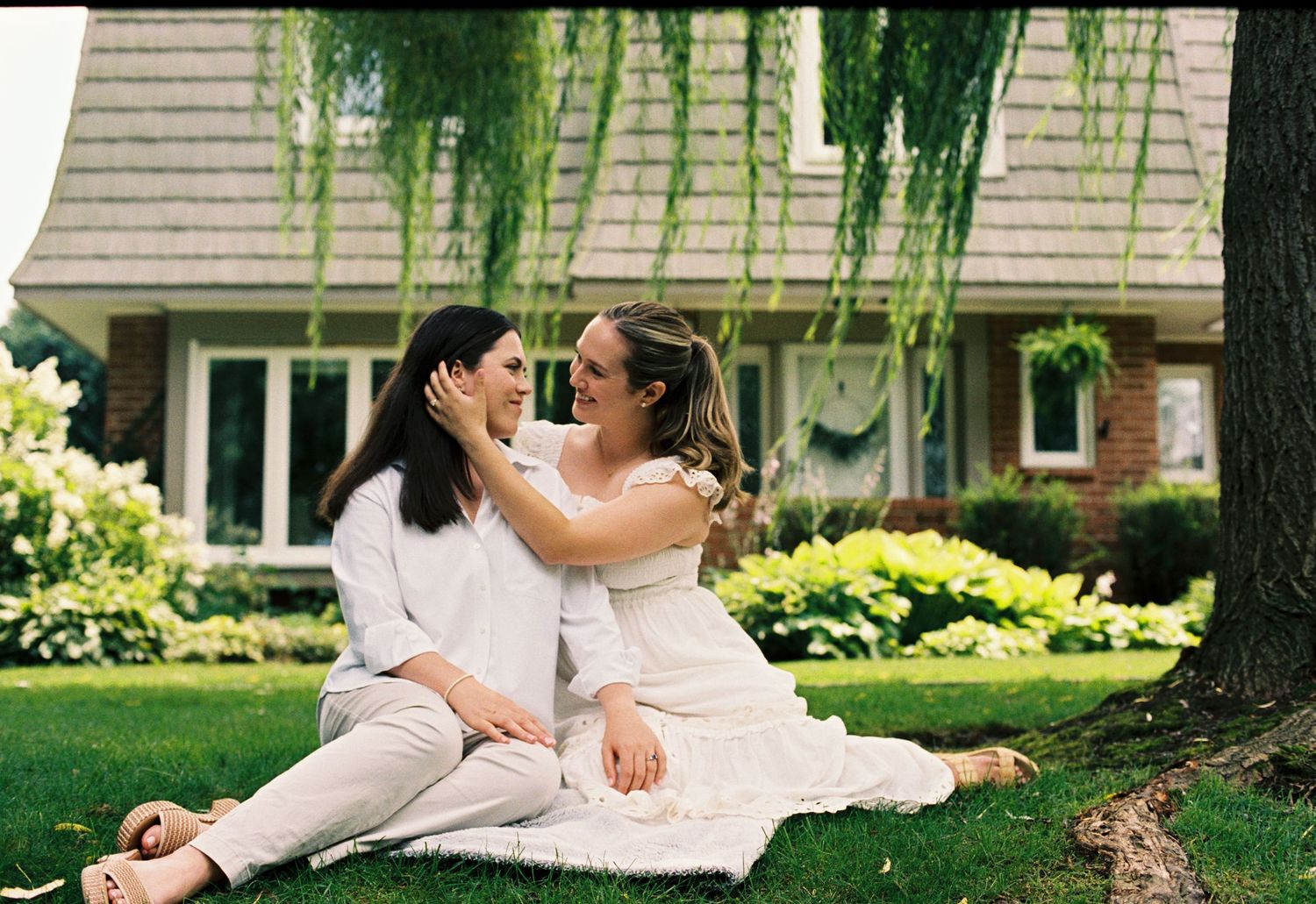 Two women in white outfits sit together on grass under a willow tree, sharing a tender moment outside a brick home.
